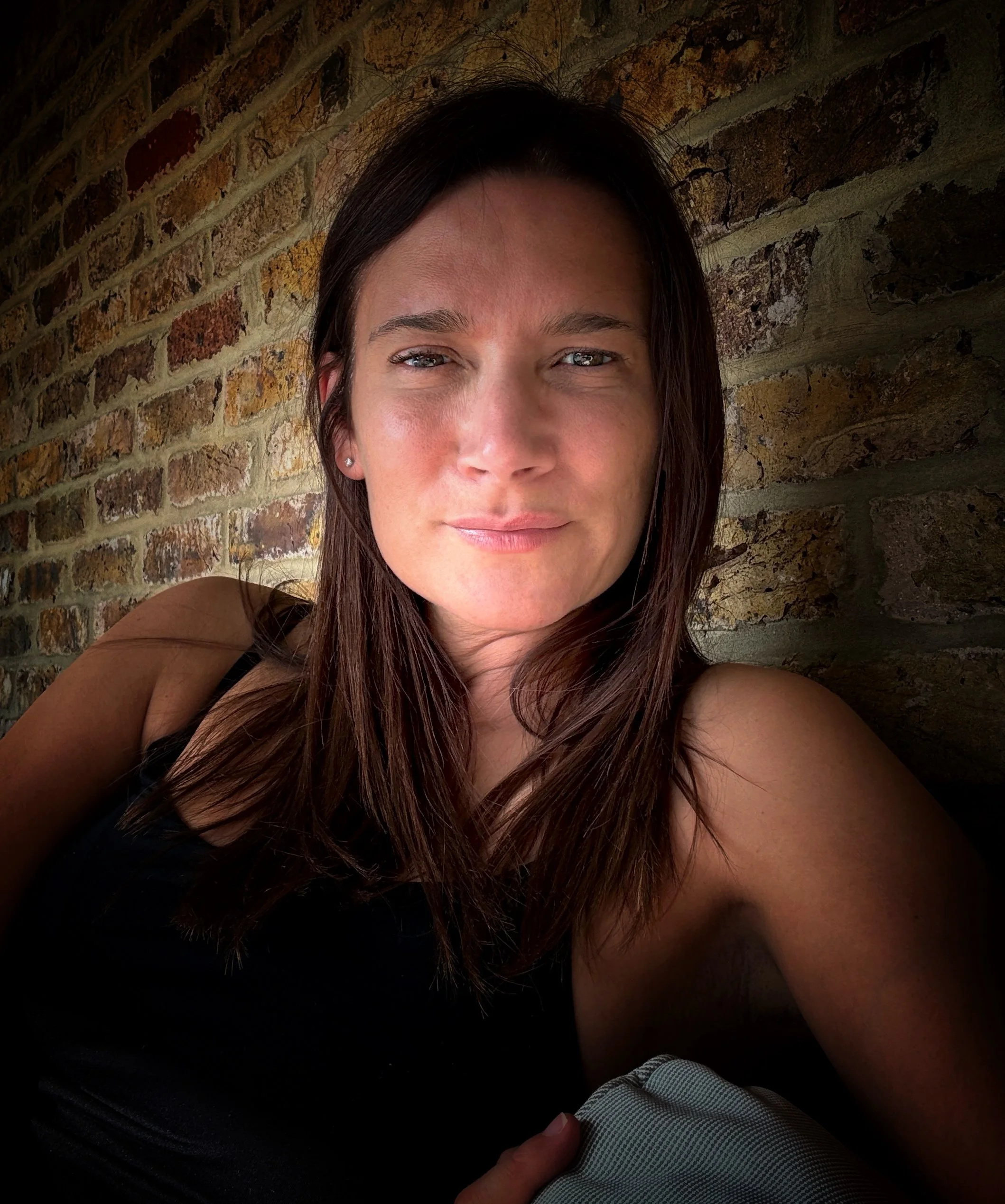 Close-up of a woman with long brown hair, blue eyes, and earrings, smiling slightly, sitting against a brick wall.