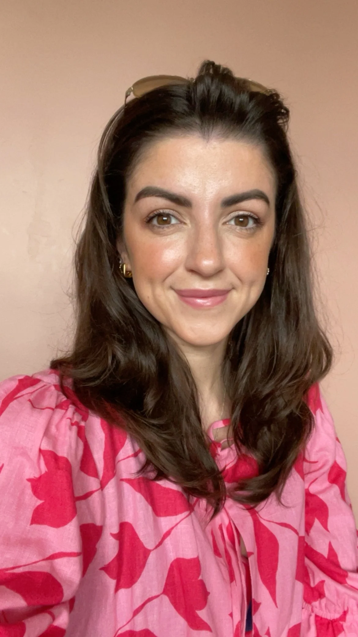 A woman with long brown hair, wearing a pink patterned blouse, smiling and looking at the camera, against a plain light pink background.