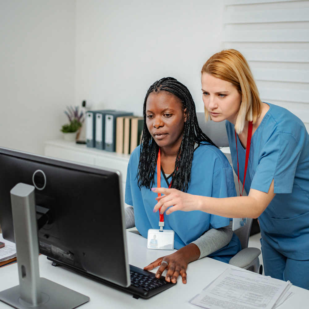 Two female healthcare workers in scrubs working together at a computer in an office.