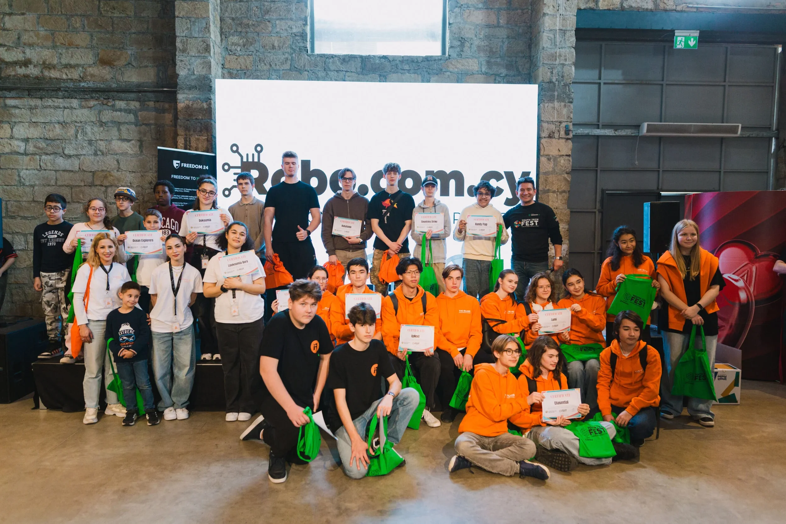 Group of young students and adults at a technology event holding certificates and green bags, standing on a stage with a large screen in the background.