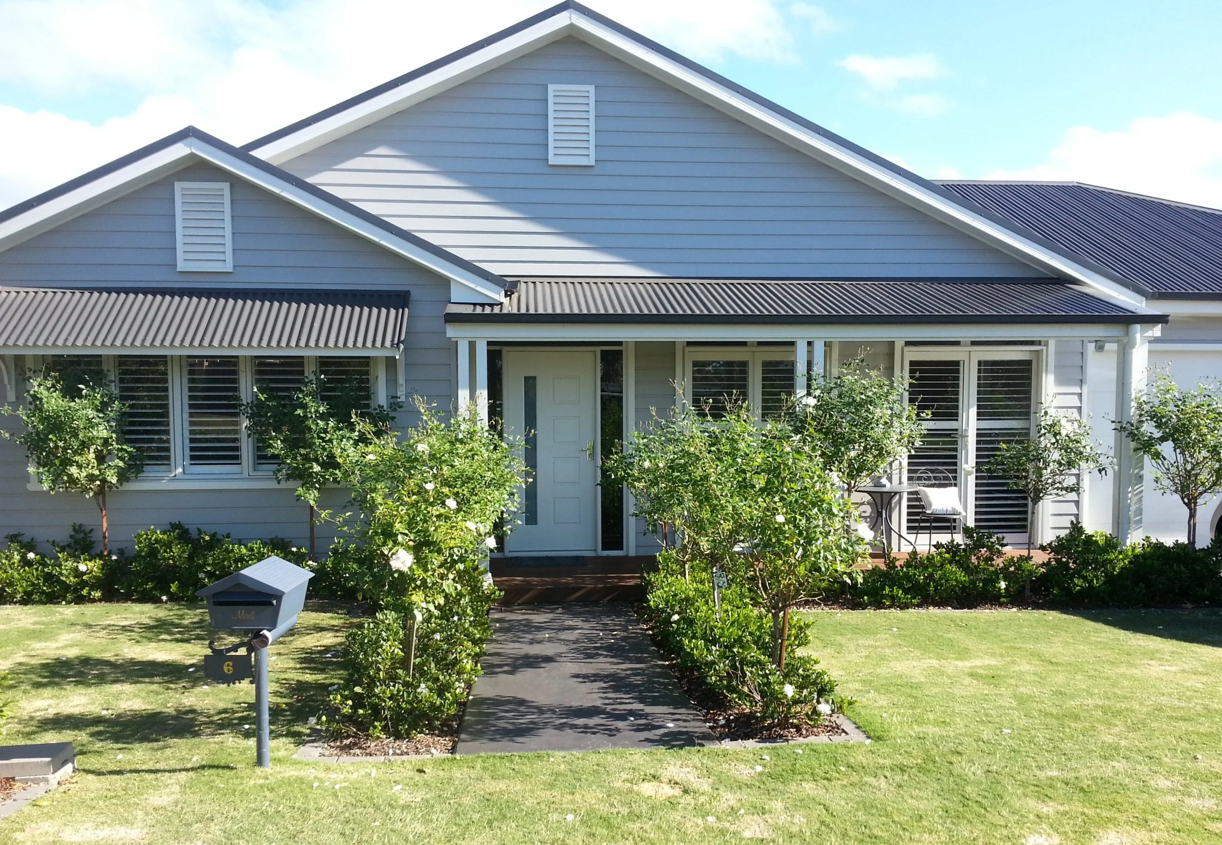 Front of a house with grey siding, white trim, a porch with seating, well-maintained garden, and a lawn leading to a street mailbox.