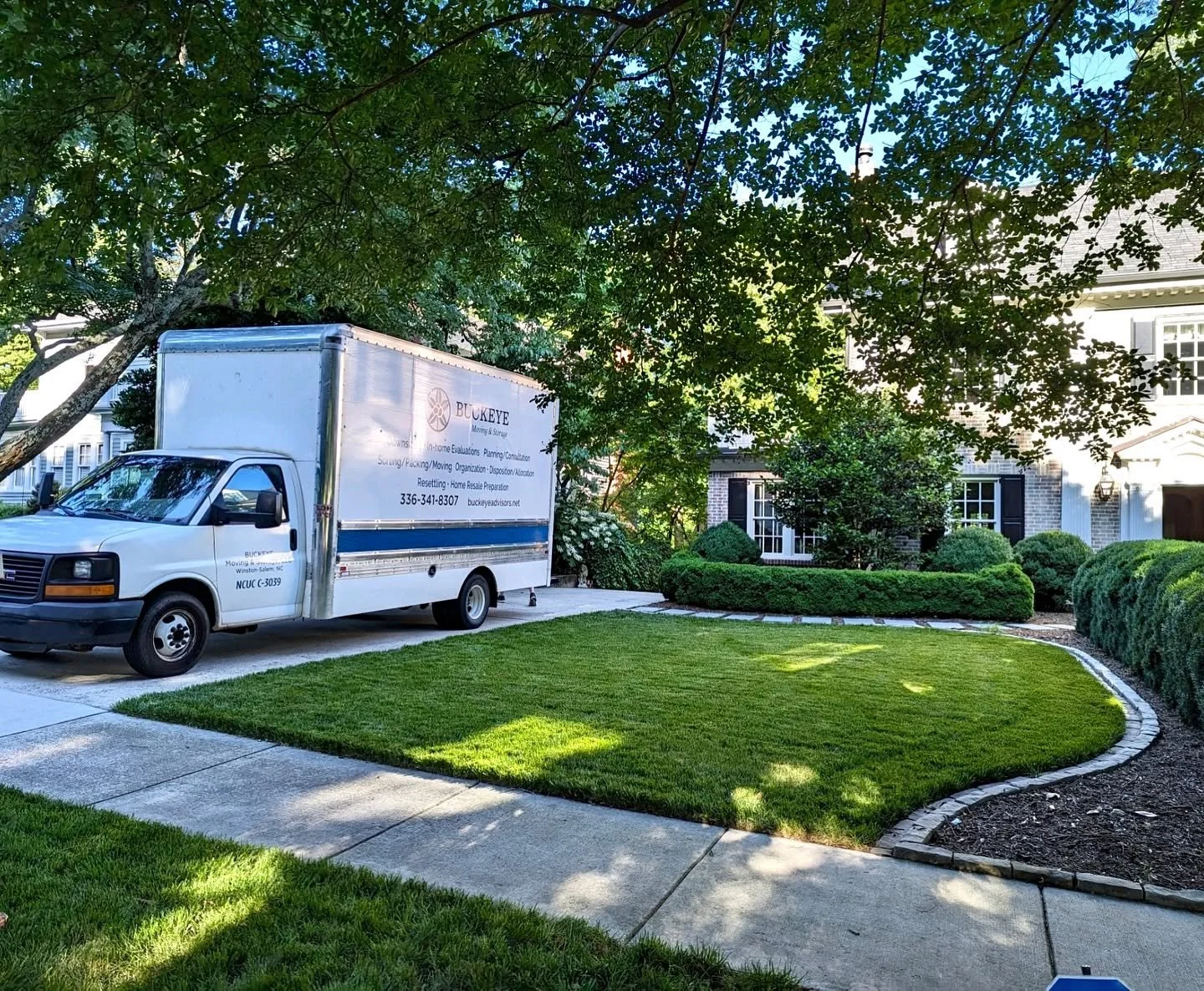 A white moving truck parked on a driveway in front of a large house with a well-manicured lawn and bushes. There are trees and a pathway beside the lawn.