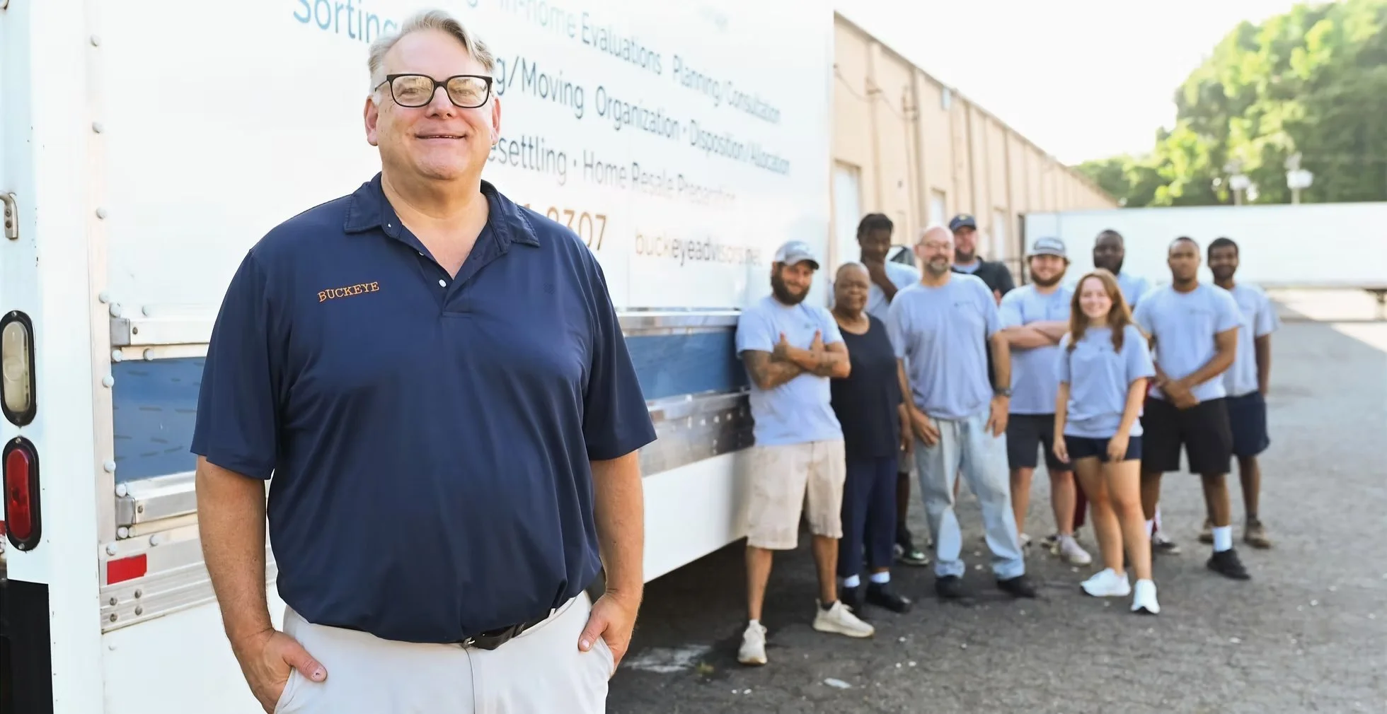 A man wearing glasses and a navy blue polo shirt with 'BUCKEYE' embroidered on it, standing in front of a group of diverse people outside near a large moving truck, with a building and trees in the background.