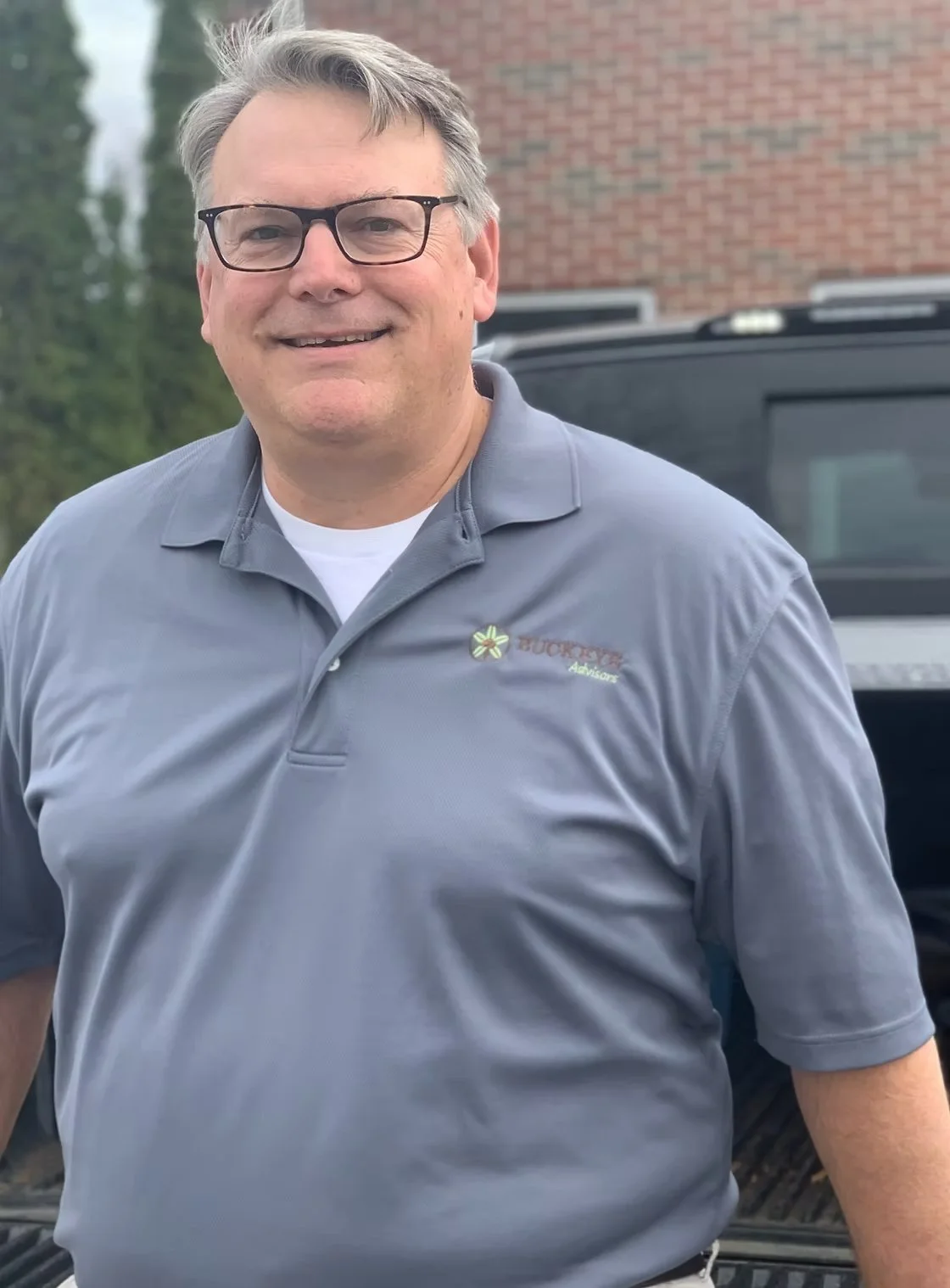 A man with glasses smiling outdoors, wearing a gray collared shirt with a logo and standing in front of a black vehicle.