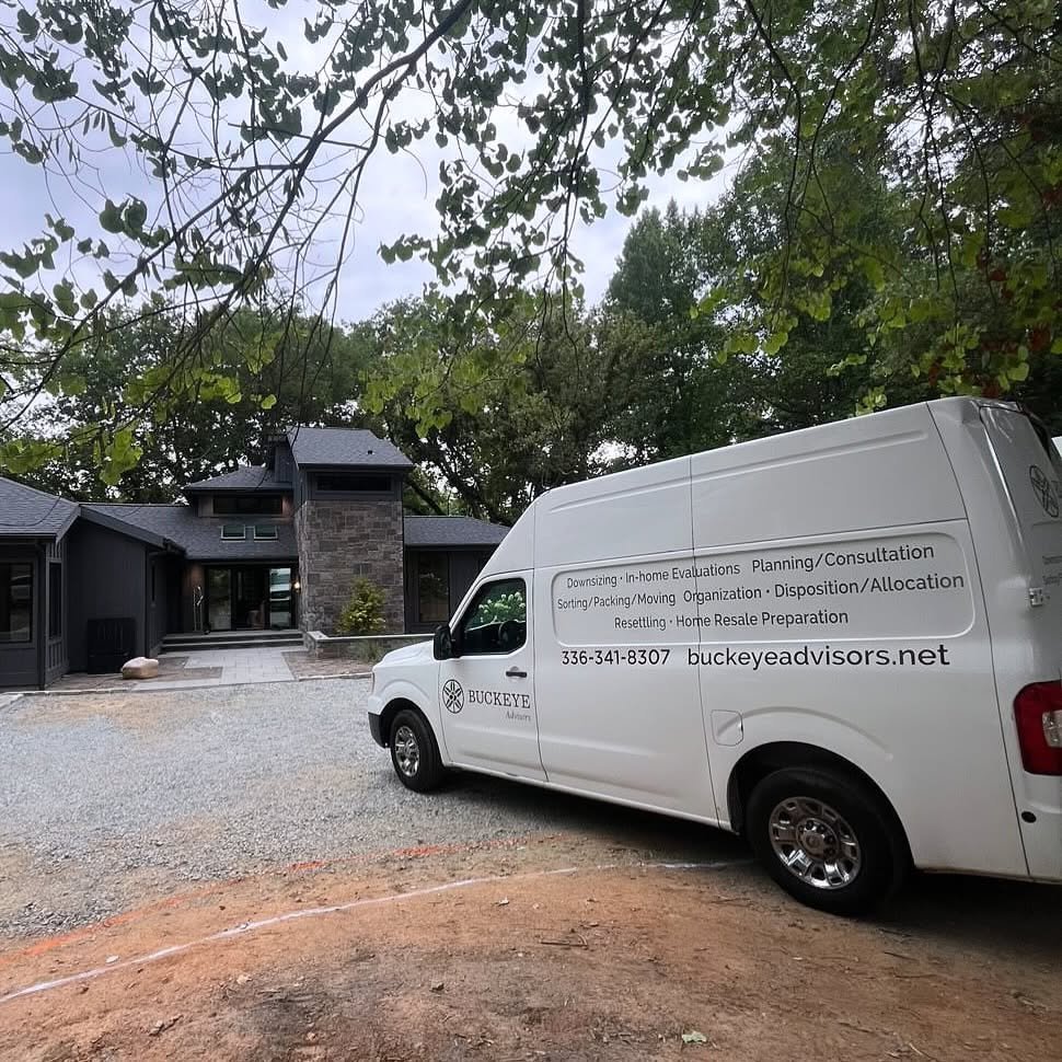 A white work van with text and a logo parked in front of a modern house with a dark exterior and stone chimney, surrounded by green trees.