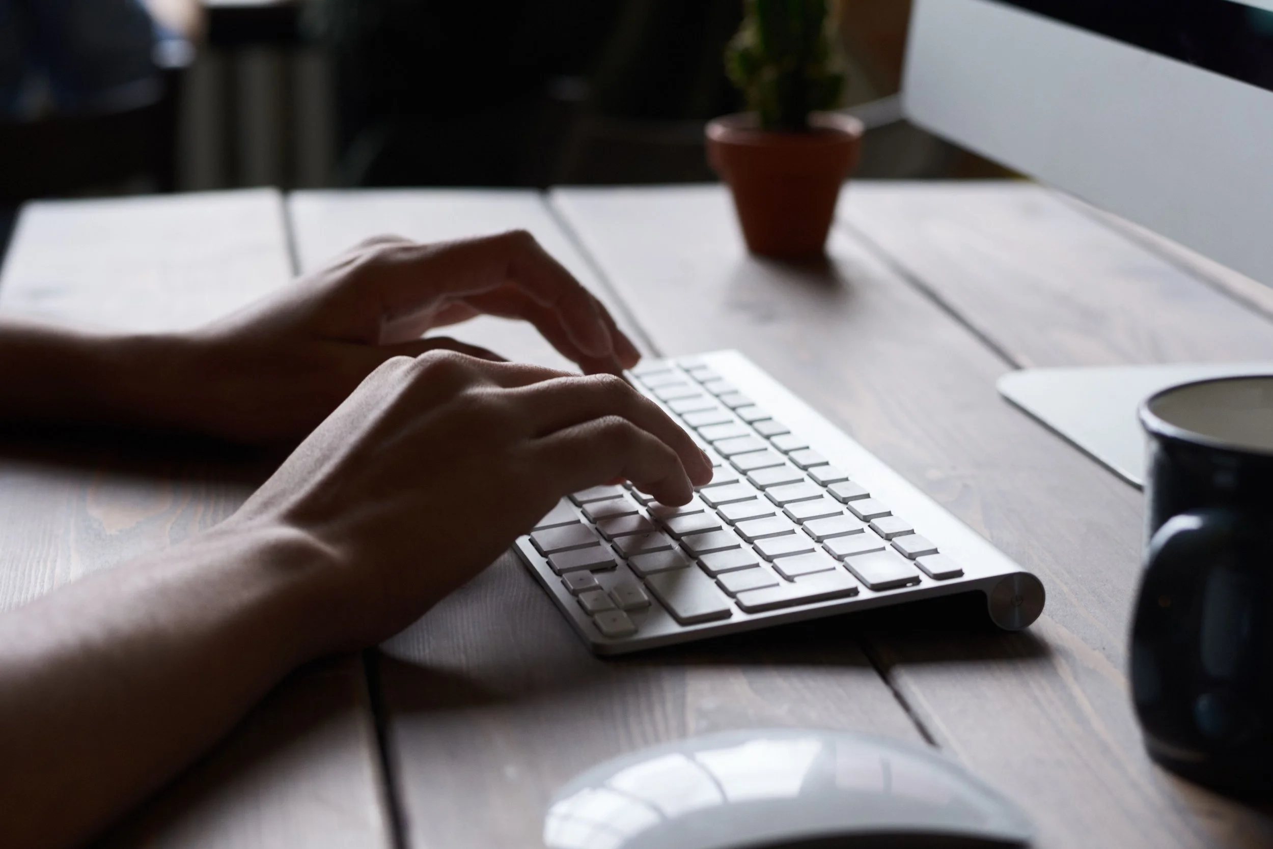 Person's hands typing on a white computer keyboard on a wooden desk, with a plant in a pot and a mug on the desk.