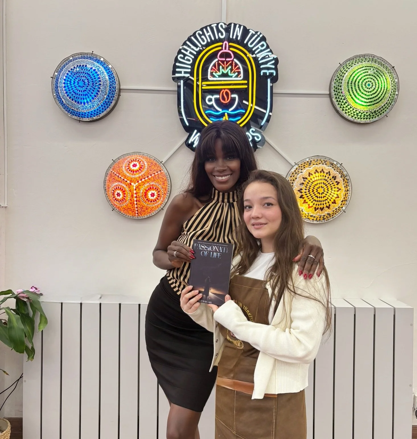 Two women smiling, standing in front of a colorful wall display with neon signs and mosaic art. The woman on the left is holding a book titled "Passionate of Life." The woman on the right is holding a brown gift bag. Plants are visible in the lower left corner.