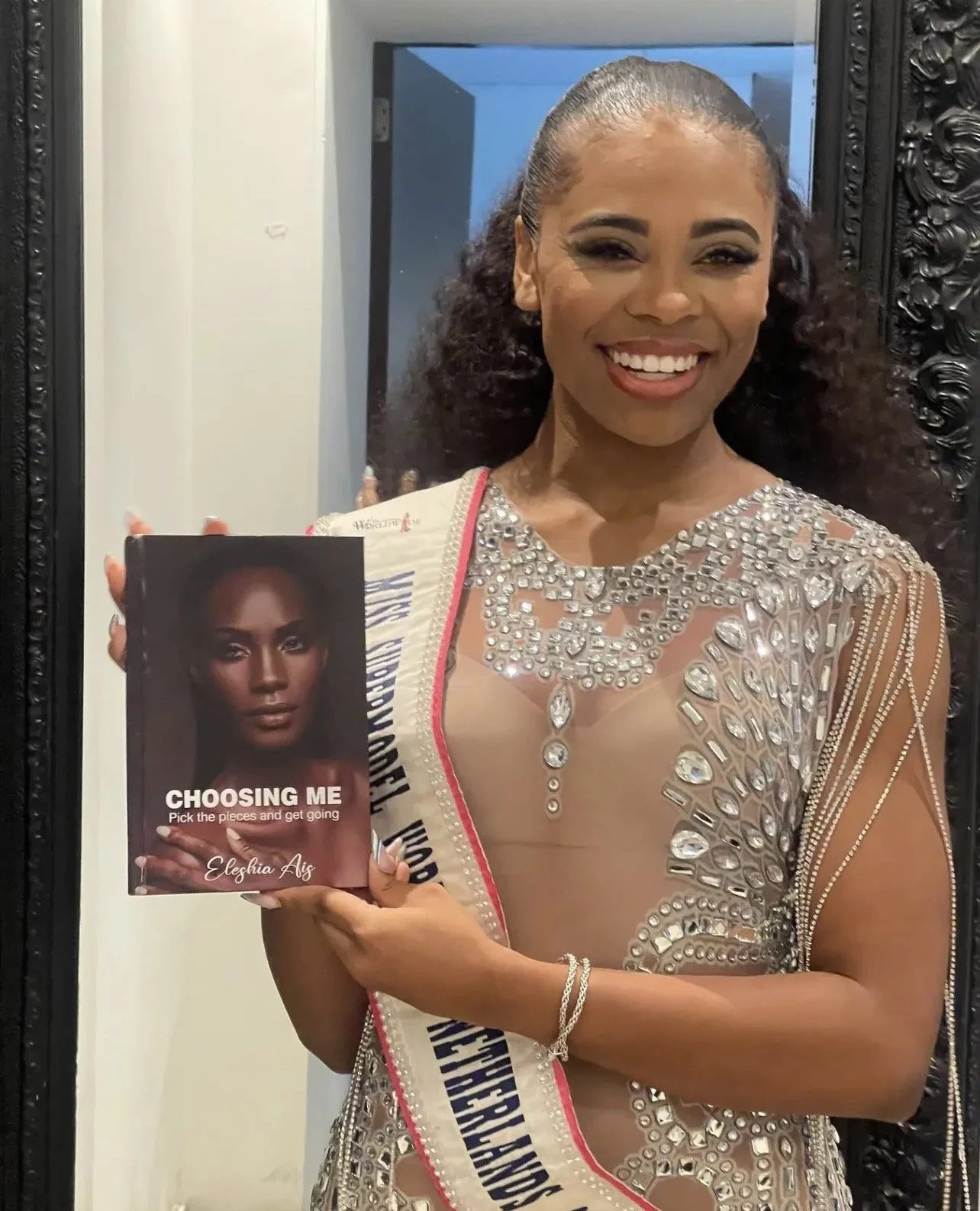 A woman in a silver dress with rhinestones, smiling while holding a book titled 'Choosing Me' by Eleshia Ais.