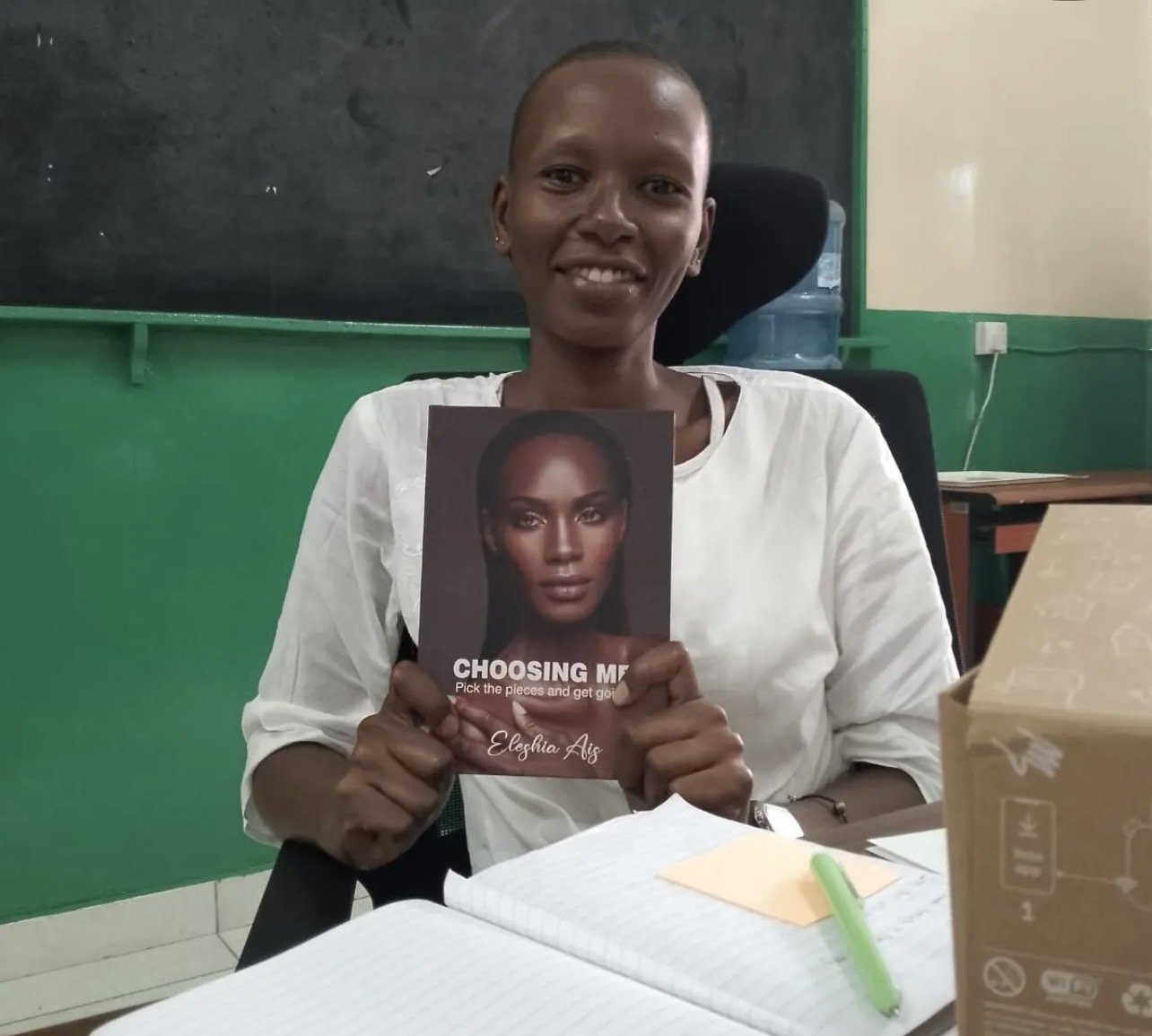 A woman sitting at a desk in a classroom, holding a book titled "Choosing Me"  by Eleshia Ais.