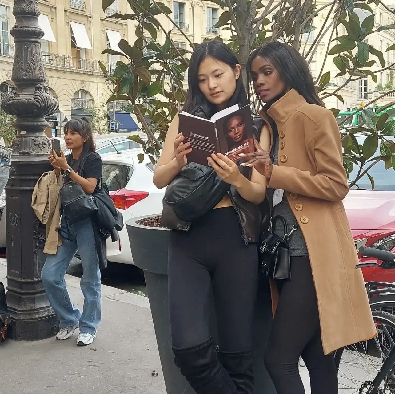 Two women stand close together, reading a booklet titled 'Choosing Me' on a city sidewalk, with parked cars and a building with balconies in the background. A third woman stands nearby, looking at her phone.
