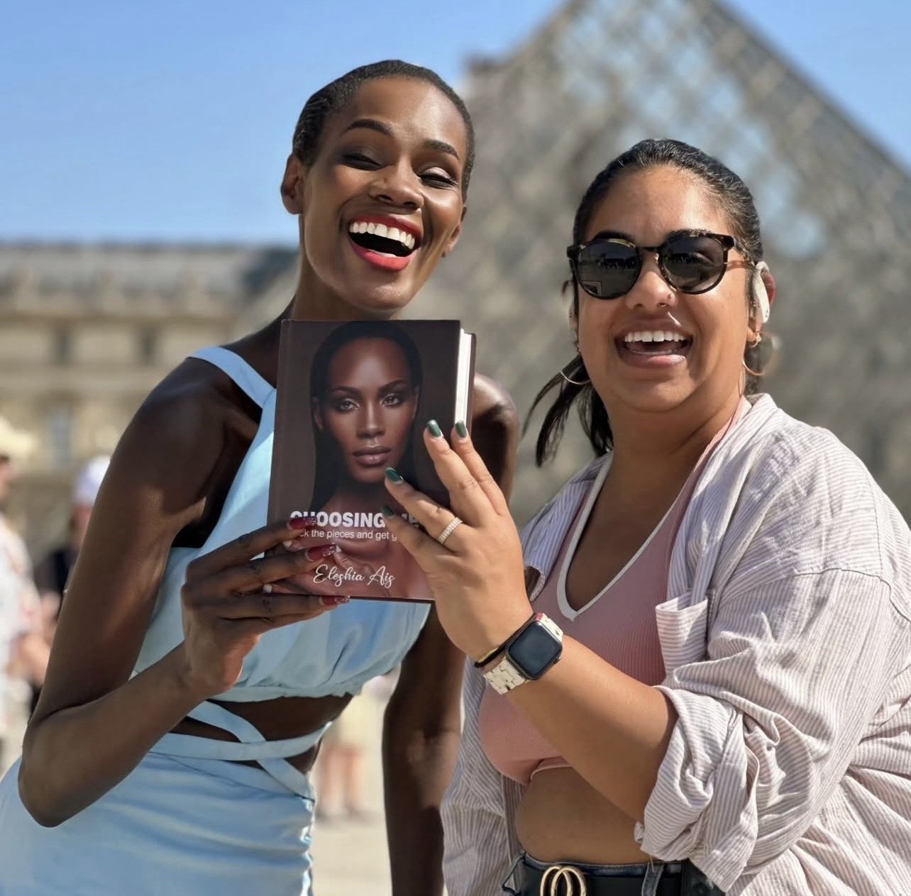 A woman  smiling after receiving a signed coppy of 'Choosing Me' book by Eleshia Ais. They are outdoors with a historic monument in the background in Paris.