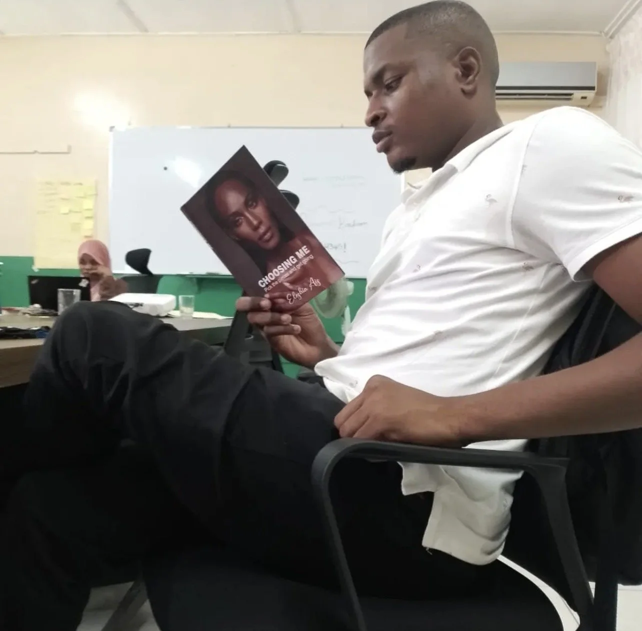 Man sitting in an office chair, reading a book  'Choosing Me' by Eleshia Ais.