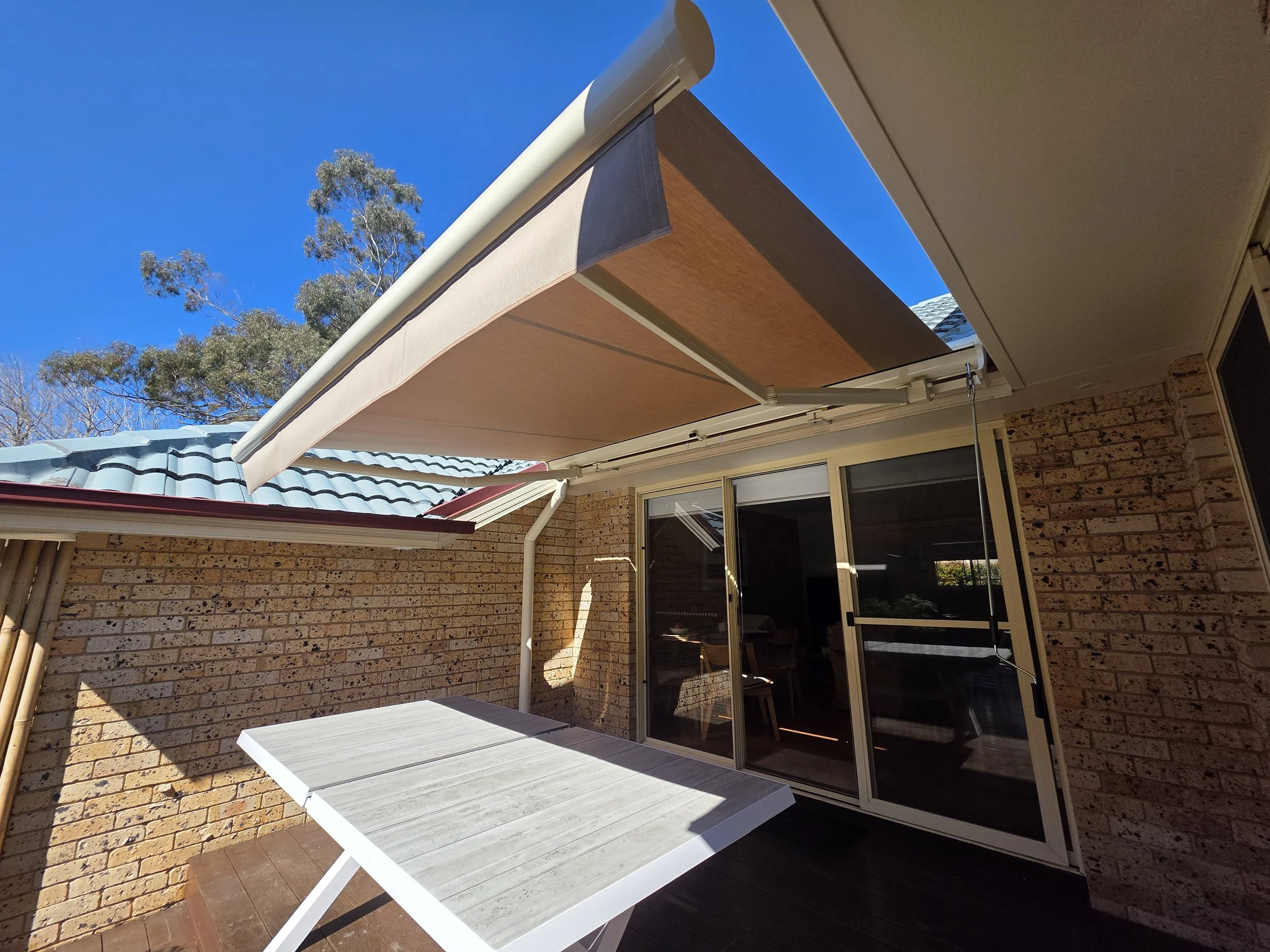 A backyard patio area with a sliding glass door, brick wall, and an outdoor table. A beige retractable awning is extended above the patio. Trees and a blue sky are visible in the background.
