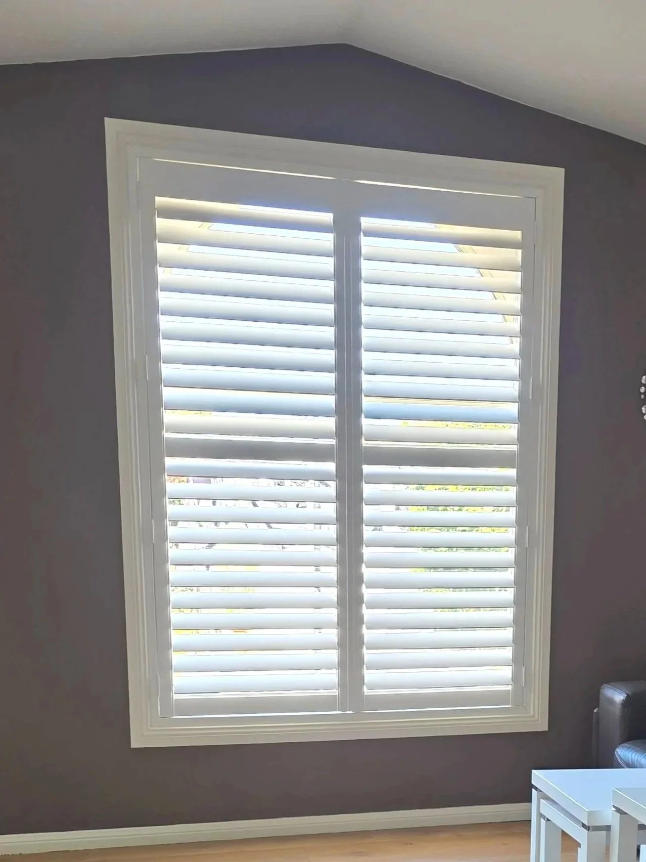 A window with white plantation shutters in a room with dark gray walls and wooden floor. Part of a white table and a chair are visible in the lower right corner.