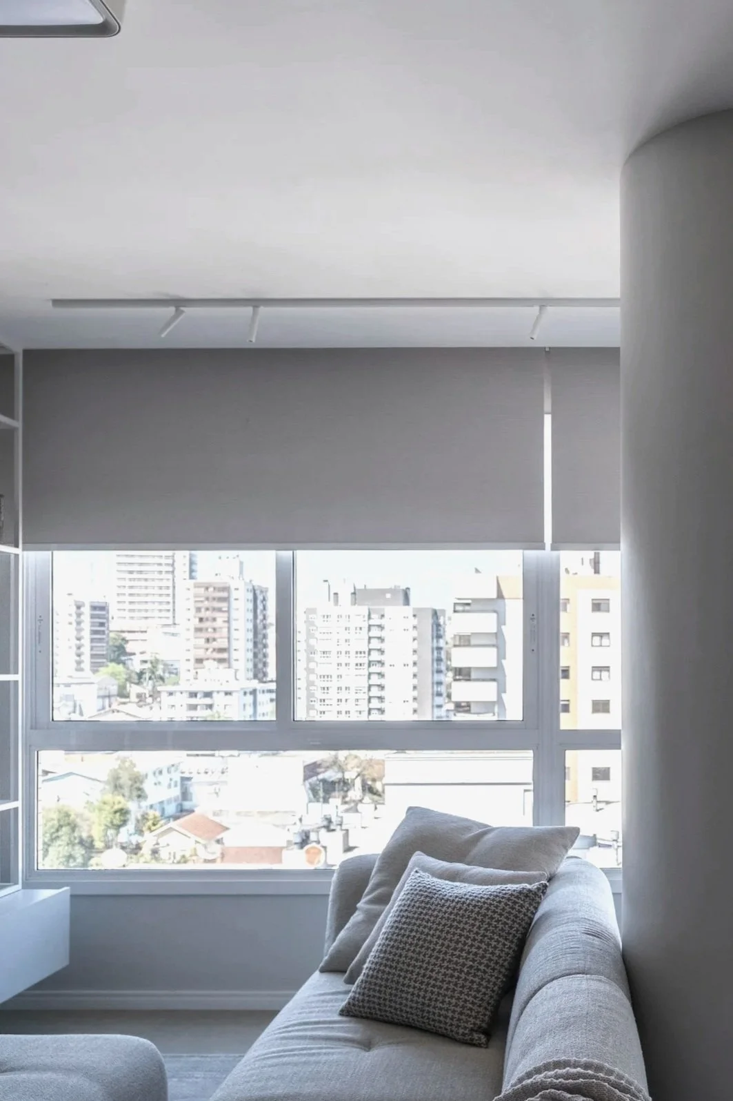 Living room with large window overlooking cityscape, grey sofa with patterned cushions, and window blinds partially closed.