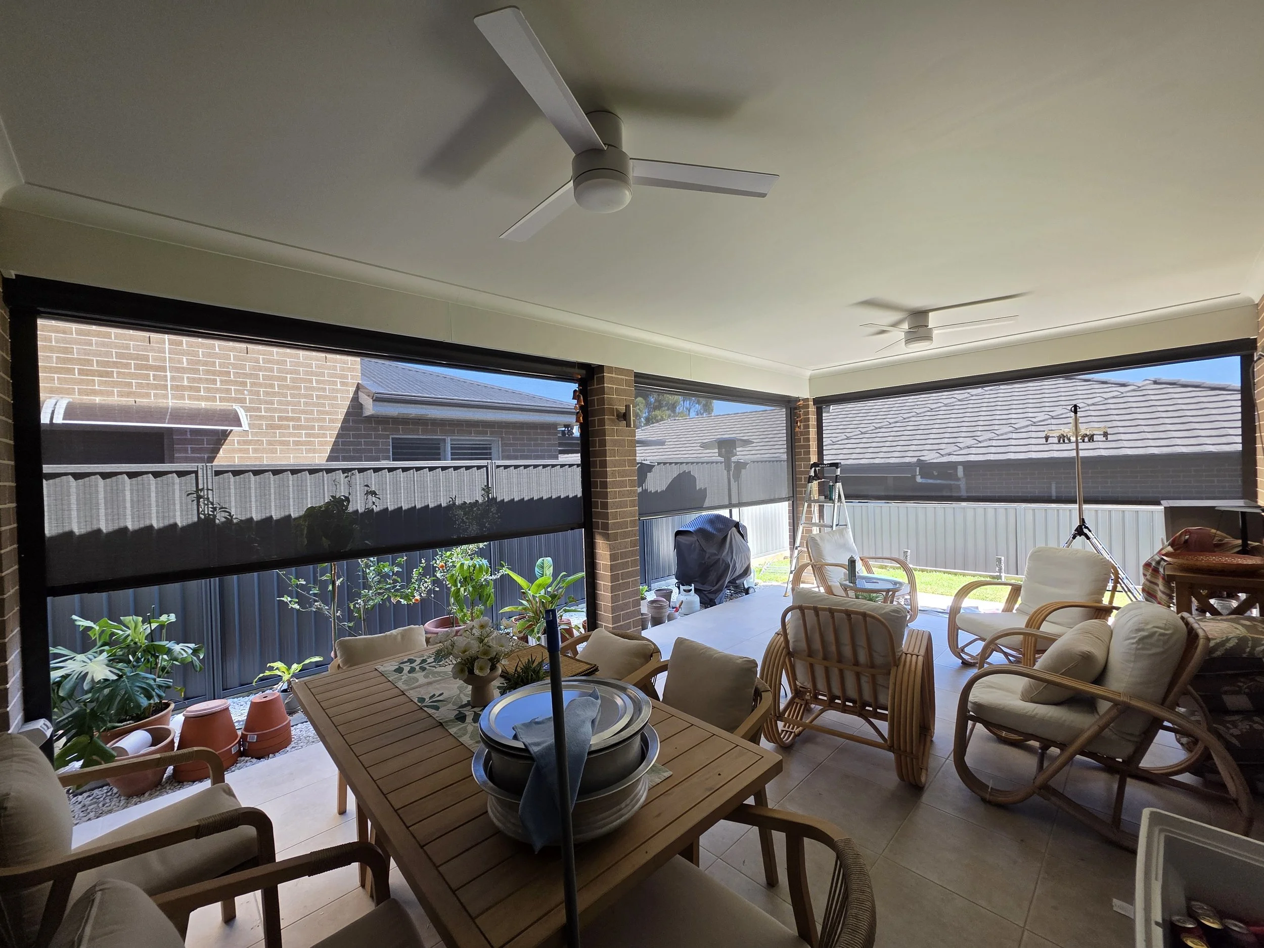 Covered backyard patio with ceiling fans, outdoor furniture, potted plants, and a view of neighboring houses through windows.