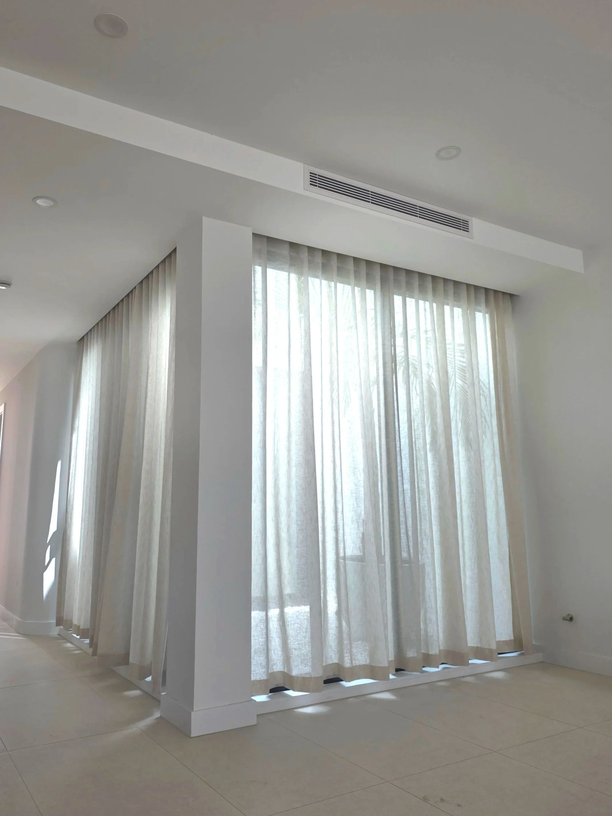 White curtains hanging in front of large windows with natural light coming through, part of an interior space.