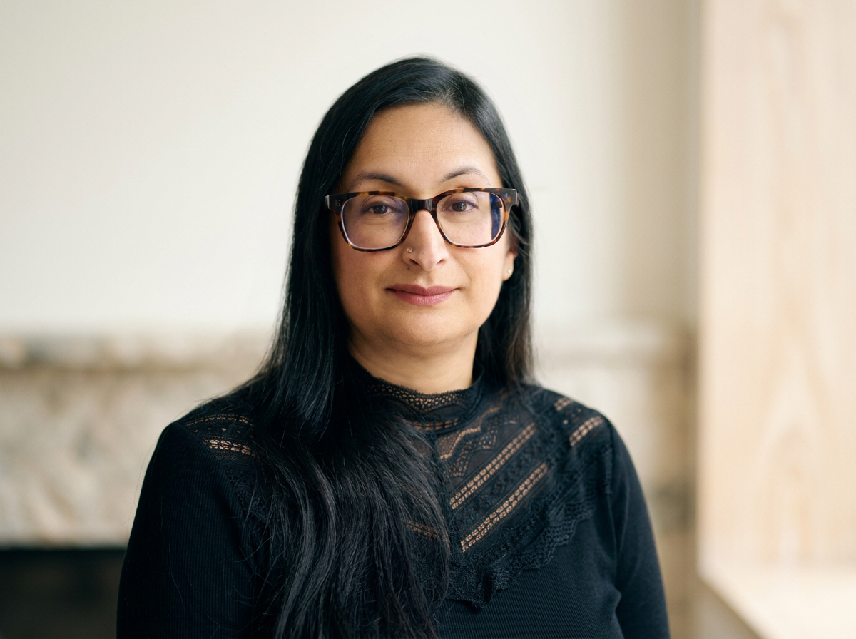 A South Asian woman with long black hair, wearing glasses, sitting indoors with a neutral background.
