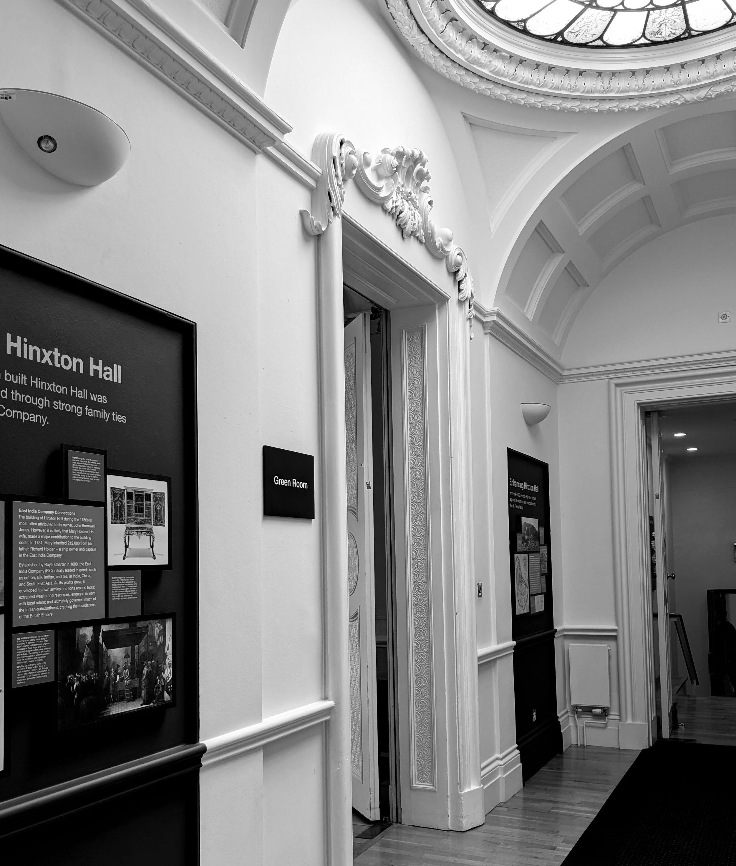 Black and white photo of an interior hallway with ornate decorative moldings near the ceiling, display boards with text and images on the walls, and a polished wooden floor.