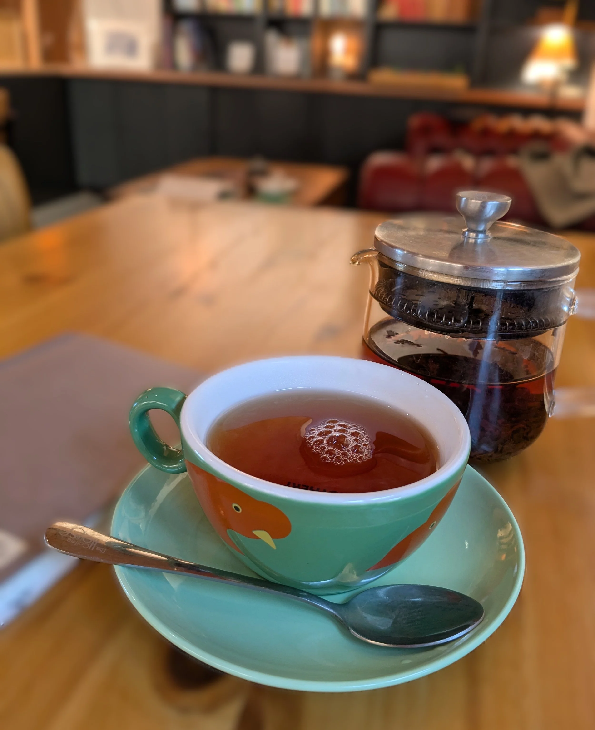 Colour photo of a large cup of tea, notebook and glass teapot, on a wooden table.