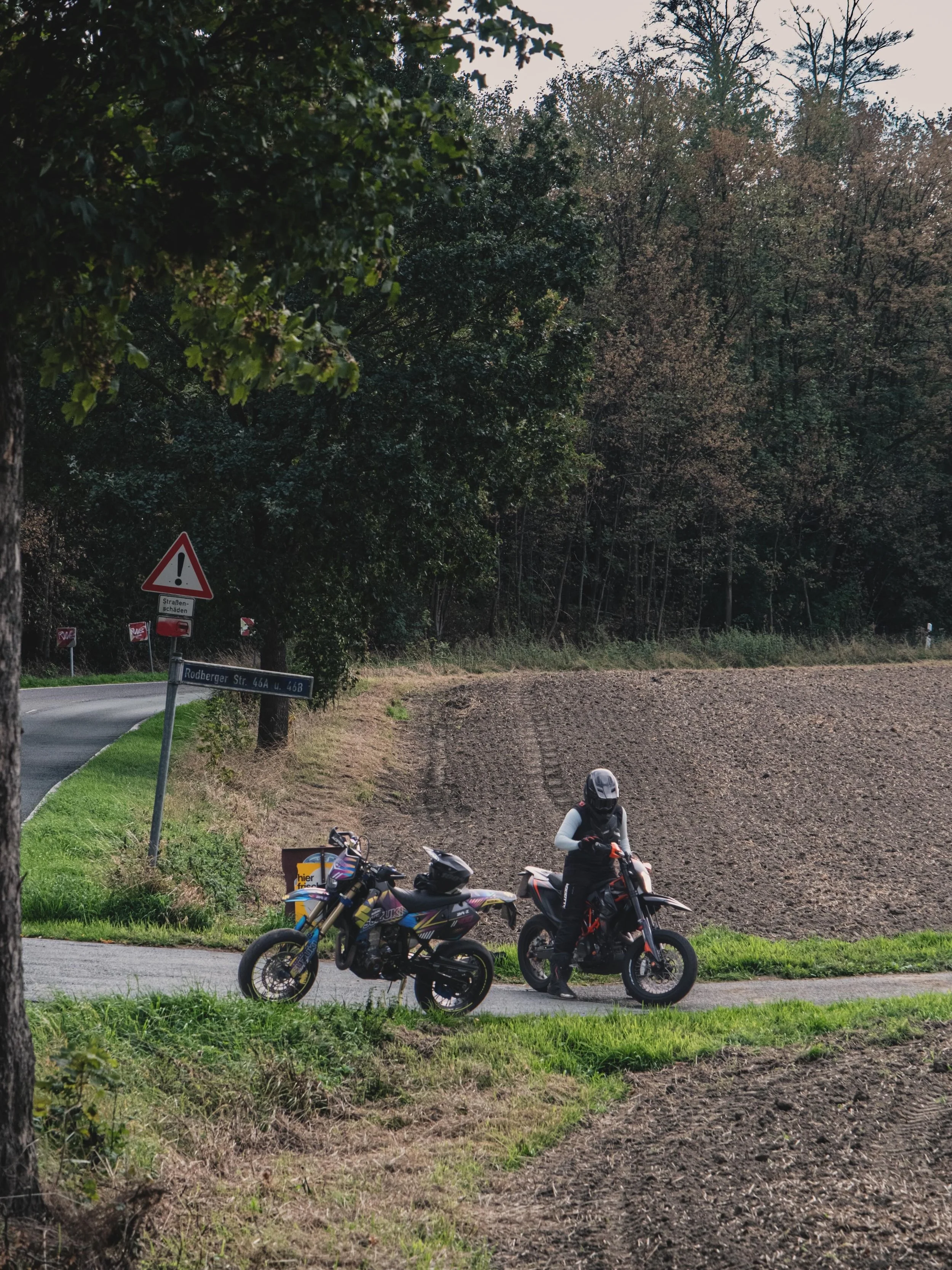 Zwei Mountainbiker stehen mit Helmen und Schutzkleidung auf einer Straße neben einer landwirtschaftlichen Fläche, im Hintergrund Bäume und ein Verkehrsschild.