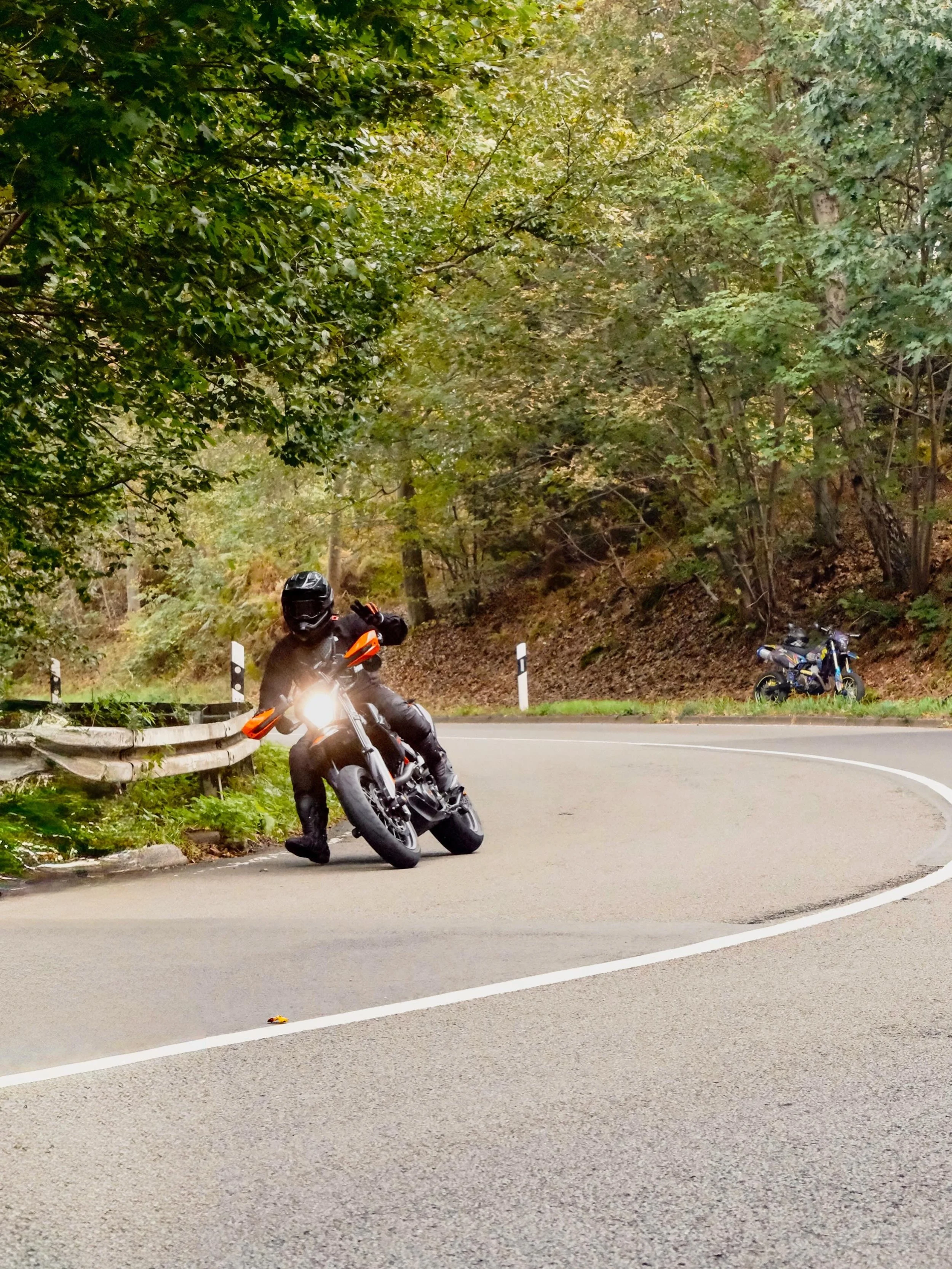 Motorradfahrer, der bei einem Kurvenausschlag auf einer Straßenstraße fährt, umgeben von Bäumen und Vegetation, mit einer Motorrad im Hintergrund