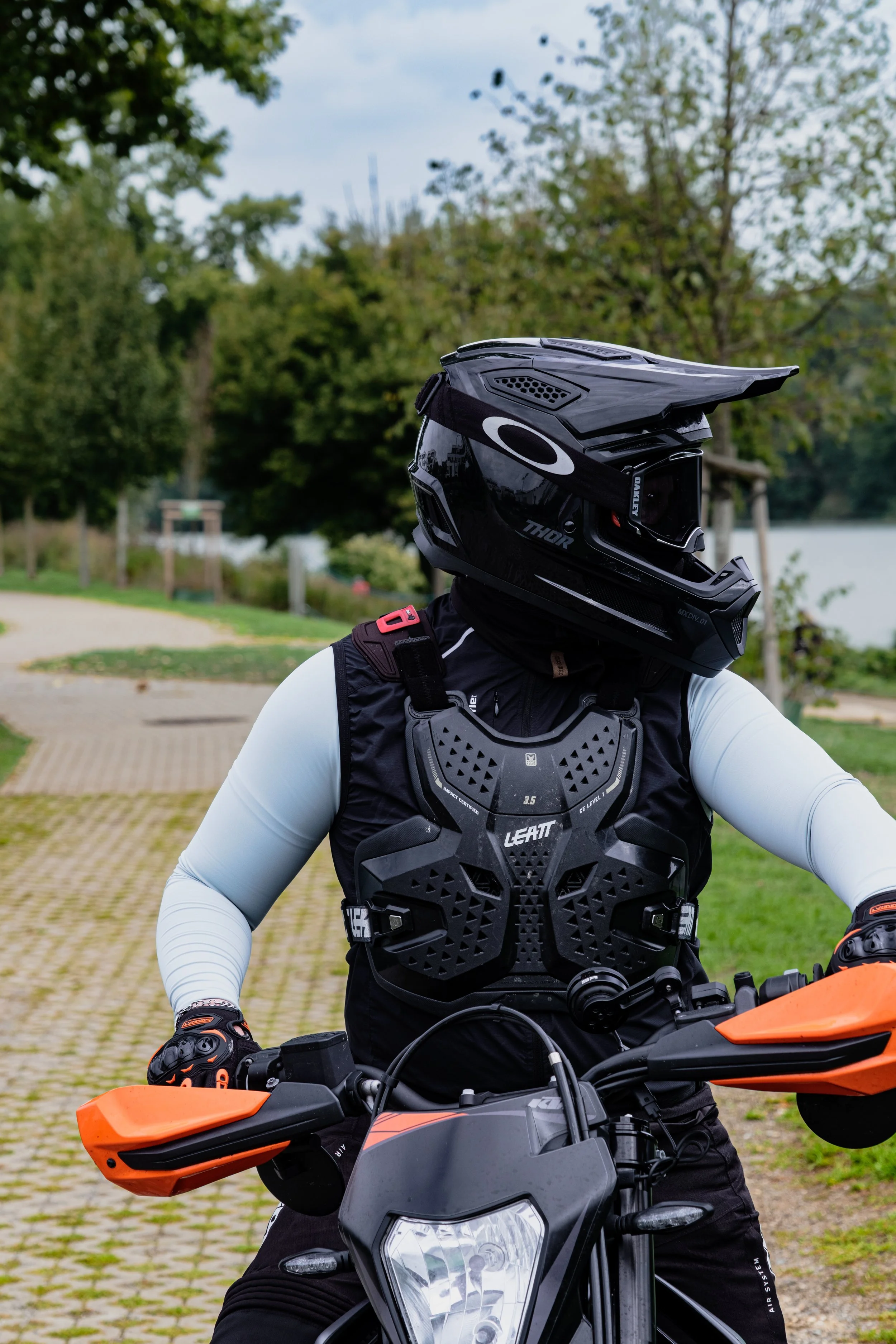 Motorradfahrer mit schwarzem Helm und Schutzweste auf orangefarbigem Motorrad im Park.