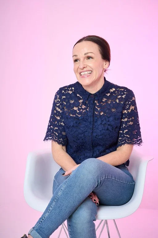 Author Elle Blair sitting on a white chair against a pink background, smiling and looking to the side, wearing a navy blue lace top and blue jeans.