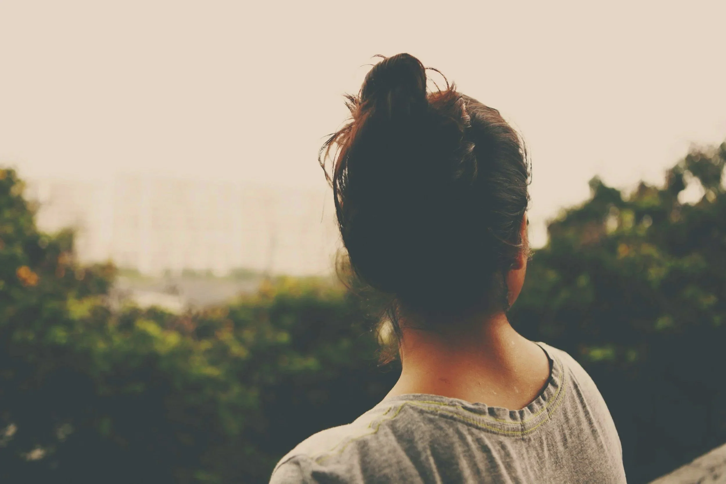 Rear view of woman with dark hair in a messy bun, wearing a striped shirt, looking into the distance outdoors with green trees and a bright sky.
