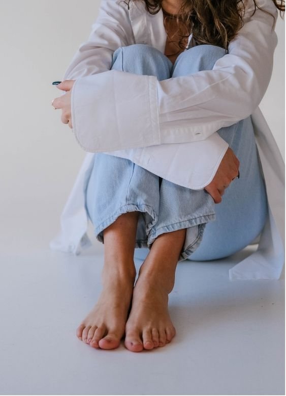 A person sitting on the floor with knees pulled to chest, wearing a white long-sleeve shirt and light blue jeans, with bare feet and toes touching the ground.