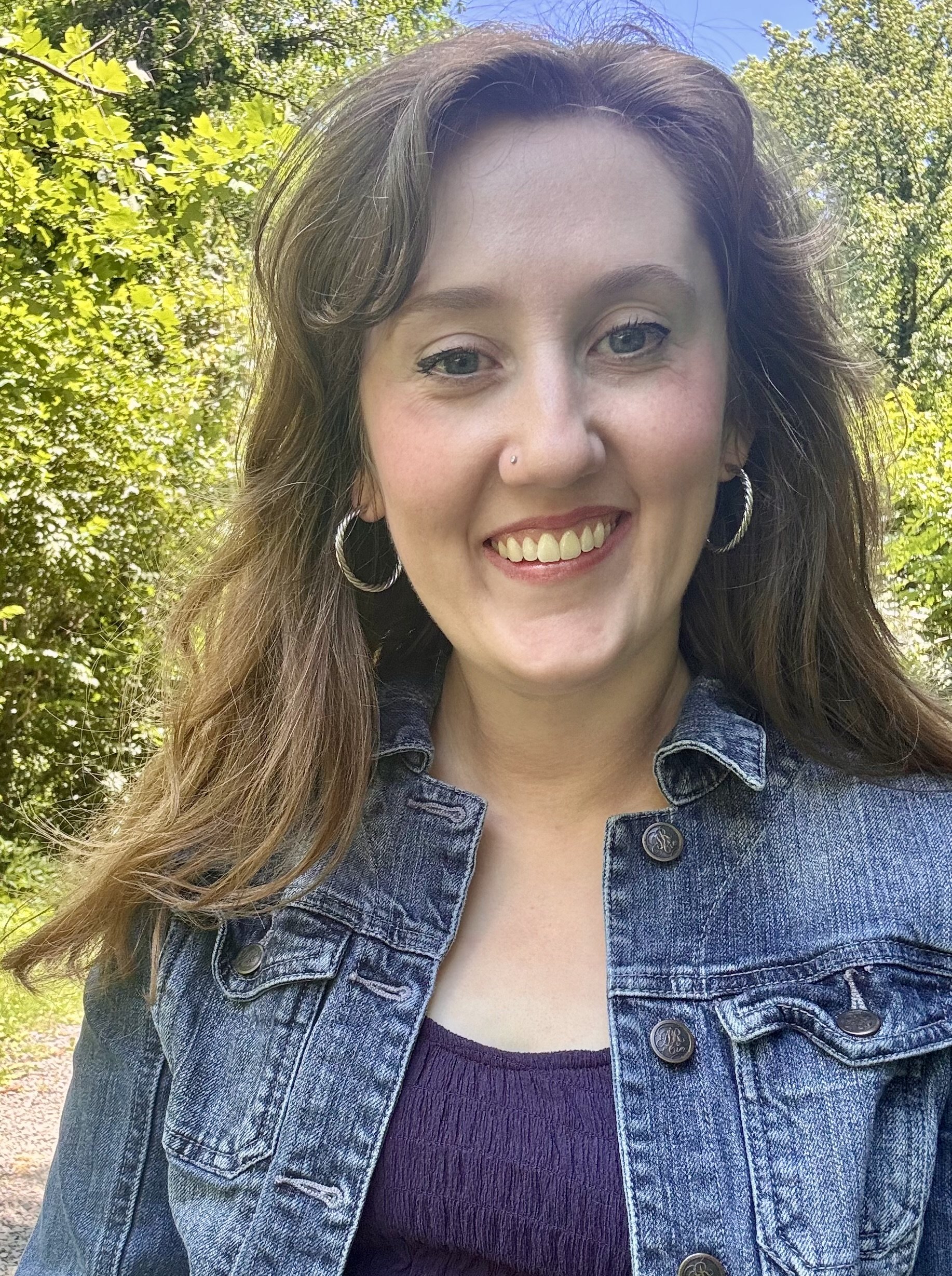 Close-up of a woman with brown hair, blue eyes, wearing hoop earrings and a denim jacket, smiling outdoors with trees and blue sky in the background.
