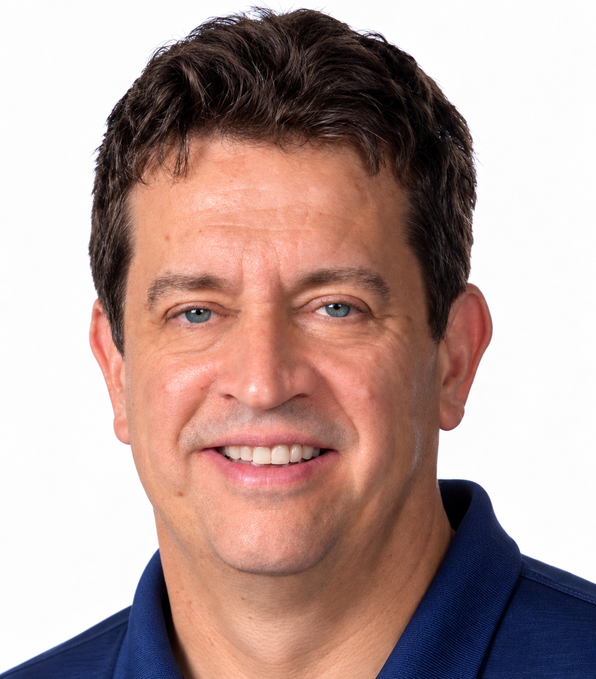 A close-up portrait of a middle-aged man with short dark hair, blue eyes, and a friendly smile, wearing a dark blue collared shirt against a white background.