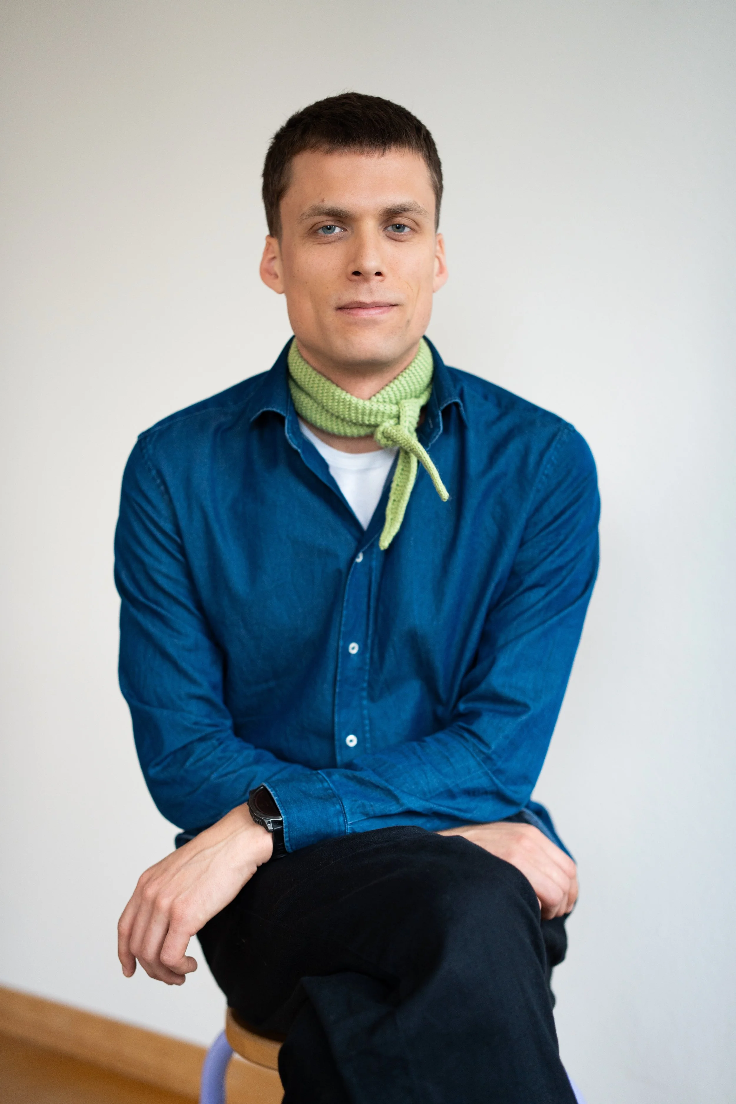 Ein junger Mann mit dunklem Haar und blauen Augen, trägt eine grüne Halskette, eine blaue Hemd und sitzt auf einem Stuhl vor einer weißen Wand