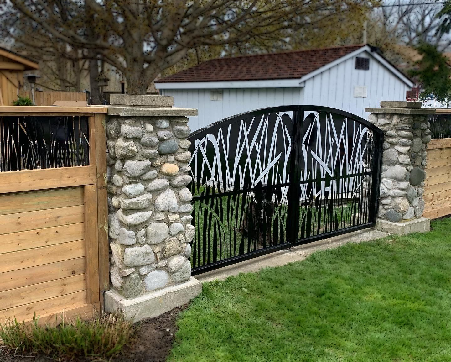 A decorative black metal gate with white streaks, flanked by stone pillars, opening to a backyard with a white shed and a tree in the background.