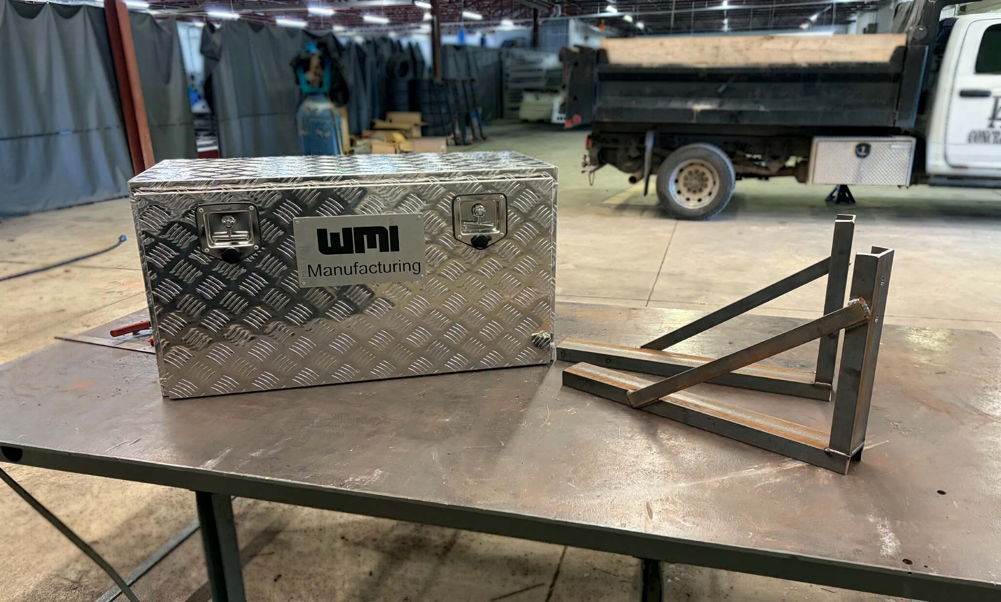 A metal box with a diamond plate pattern, labeled 'WDI Manufacturing,' is on a worktable in an industrial warehouse. In the background, there is a small truck with an open bed, various tools, equipment, and storage racks.