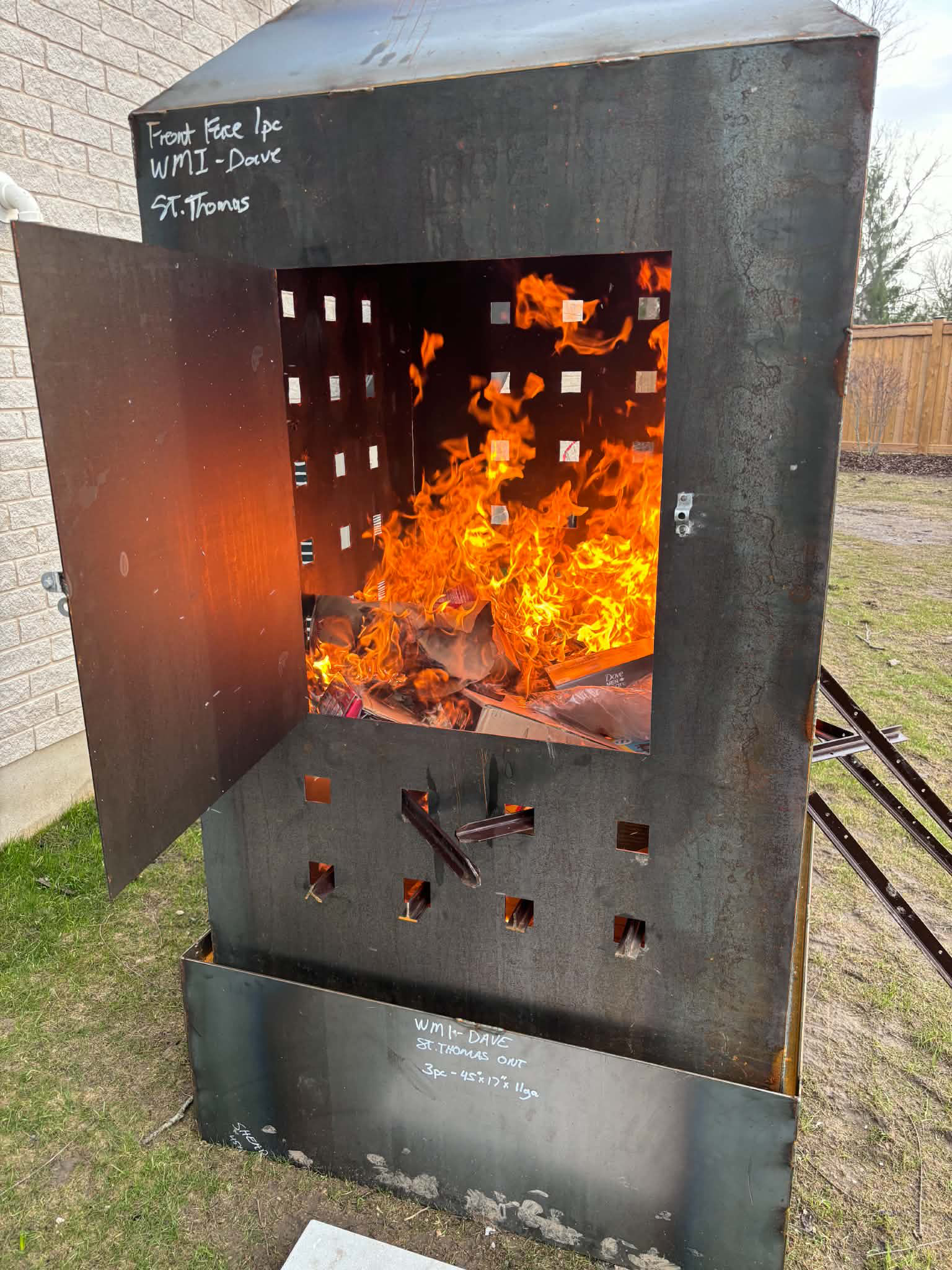 A metal barbecue smoker with flames inside. Handwritten notes on the top and bottom indicating the type of wood and wood chips used, along with other specifications.