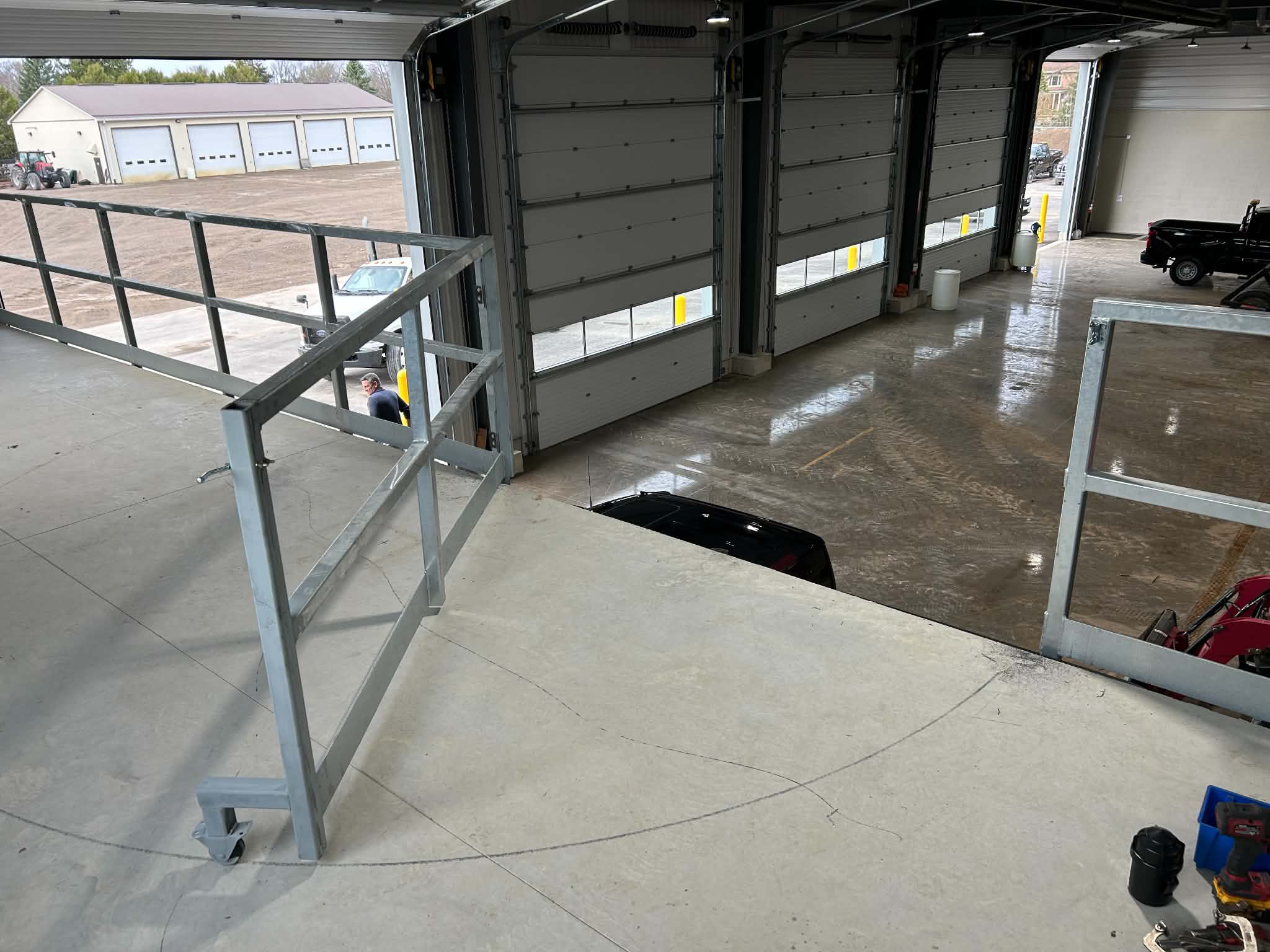Interior view of an open garage with two black garage doors, a black truck parked inside, and tools on the floor. Outside, a man is walking near a parking lot with trees and a building in the background.