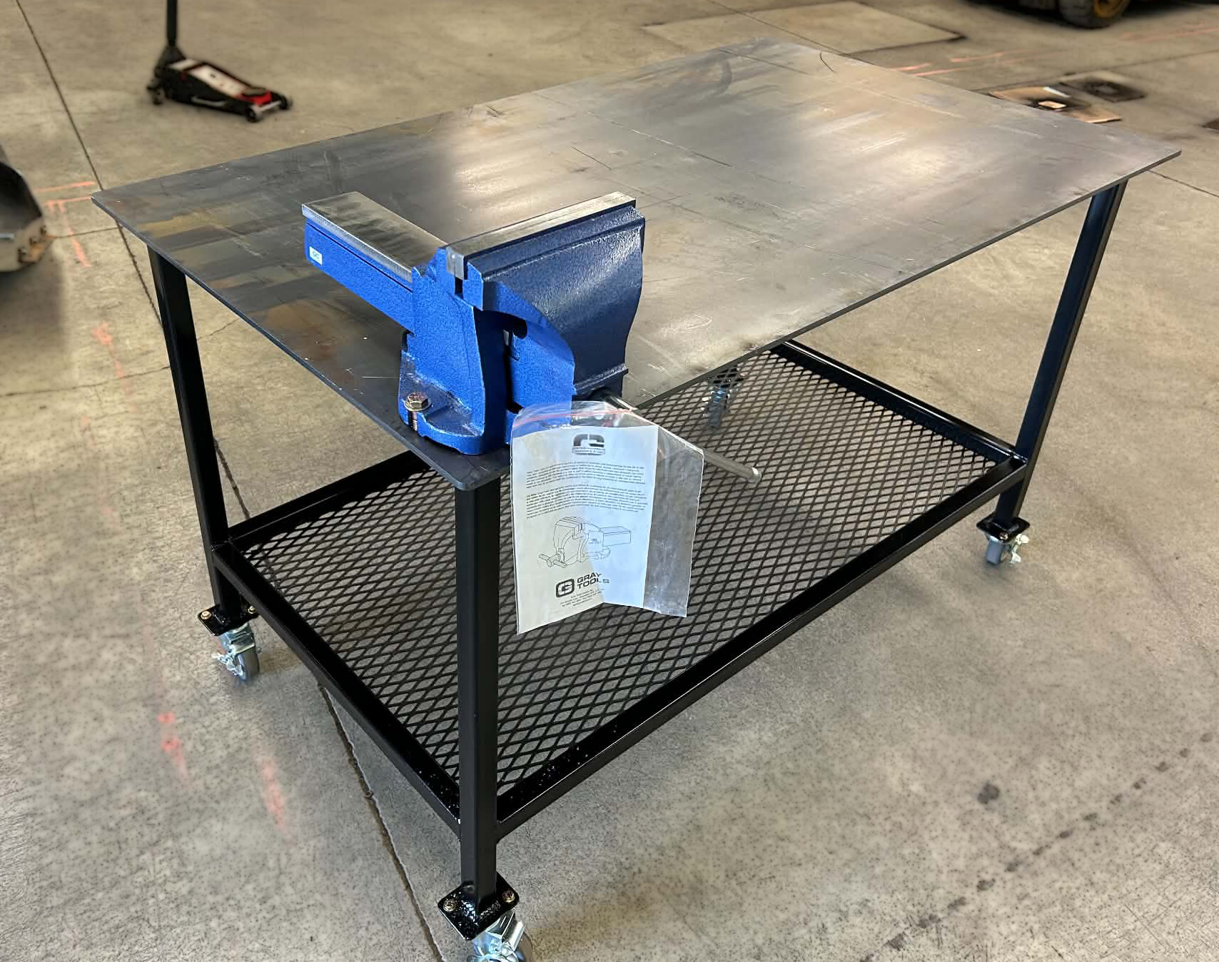 A black metal work table with a lower mesh shelf on wheels, placed on a concrete floor in an industrial setting. A blue bench vise is mounted on the left side of the table, and a plastic bag with an instruction manual is hanging from the vise.