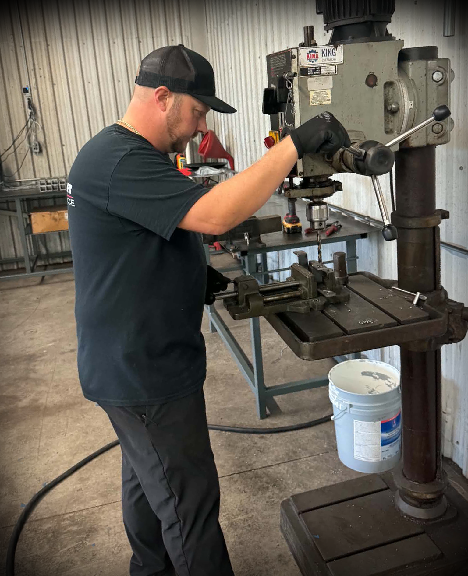 A man wearing a black cap, black gloves, and a black t-shirt working with a large industrial machine in a workshop, with a white bucket underneath.
