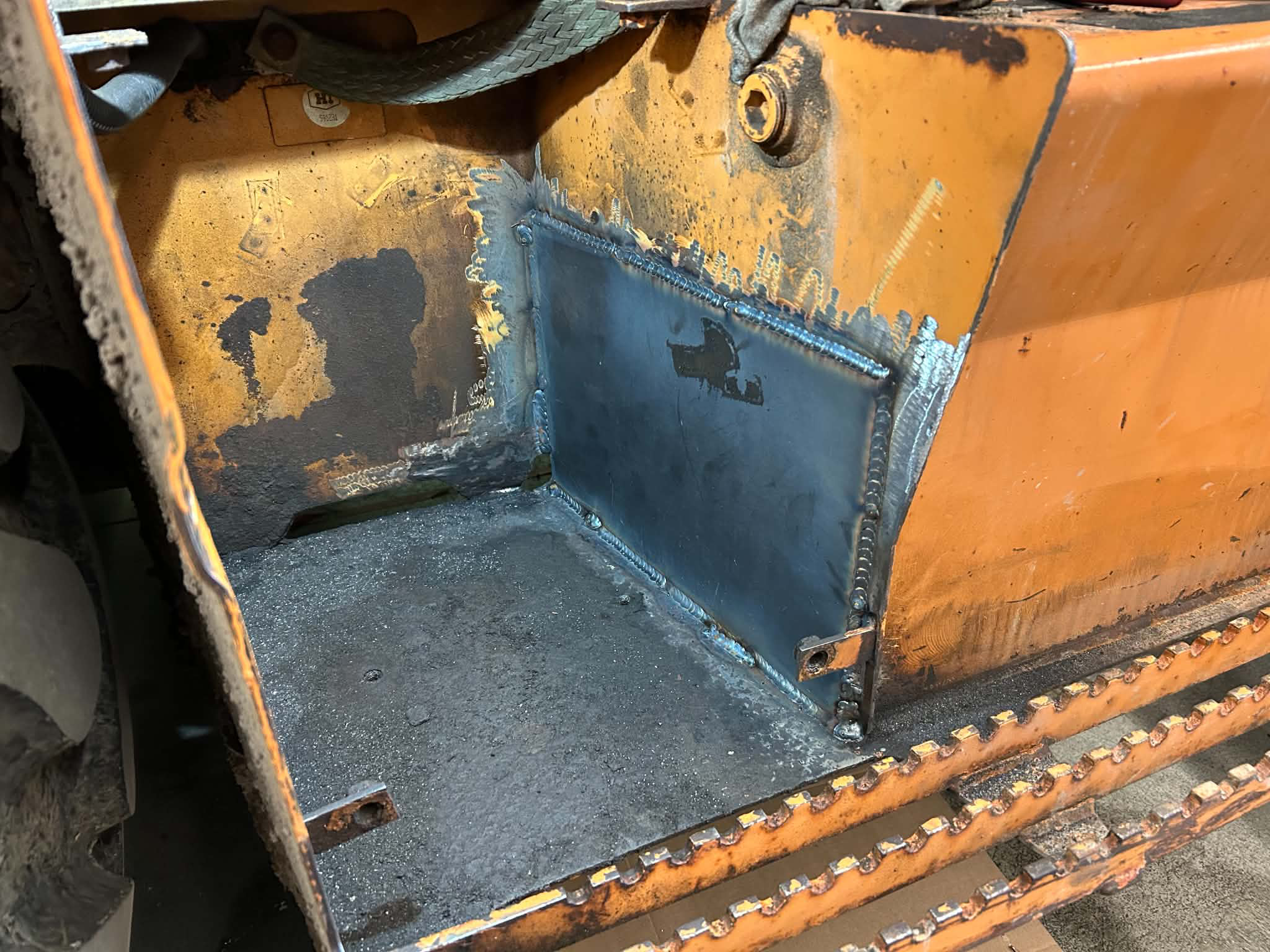 Close-up view of a welded metal box inside a construction or heavy machinery, with visible welding lines and an orange exterior. The surrounding area shows parts of the machinery with dirt and wear.