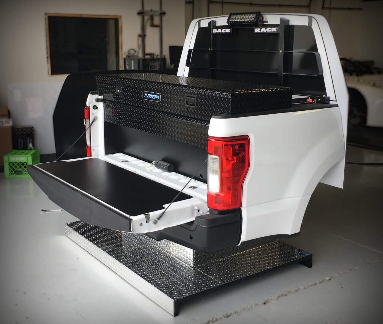 White pickup truck with a black diamond-plate toolbox in the truck bed, open tailgate, and a black diamond-plate step at the rear.