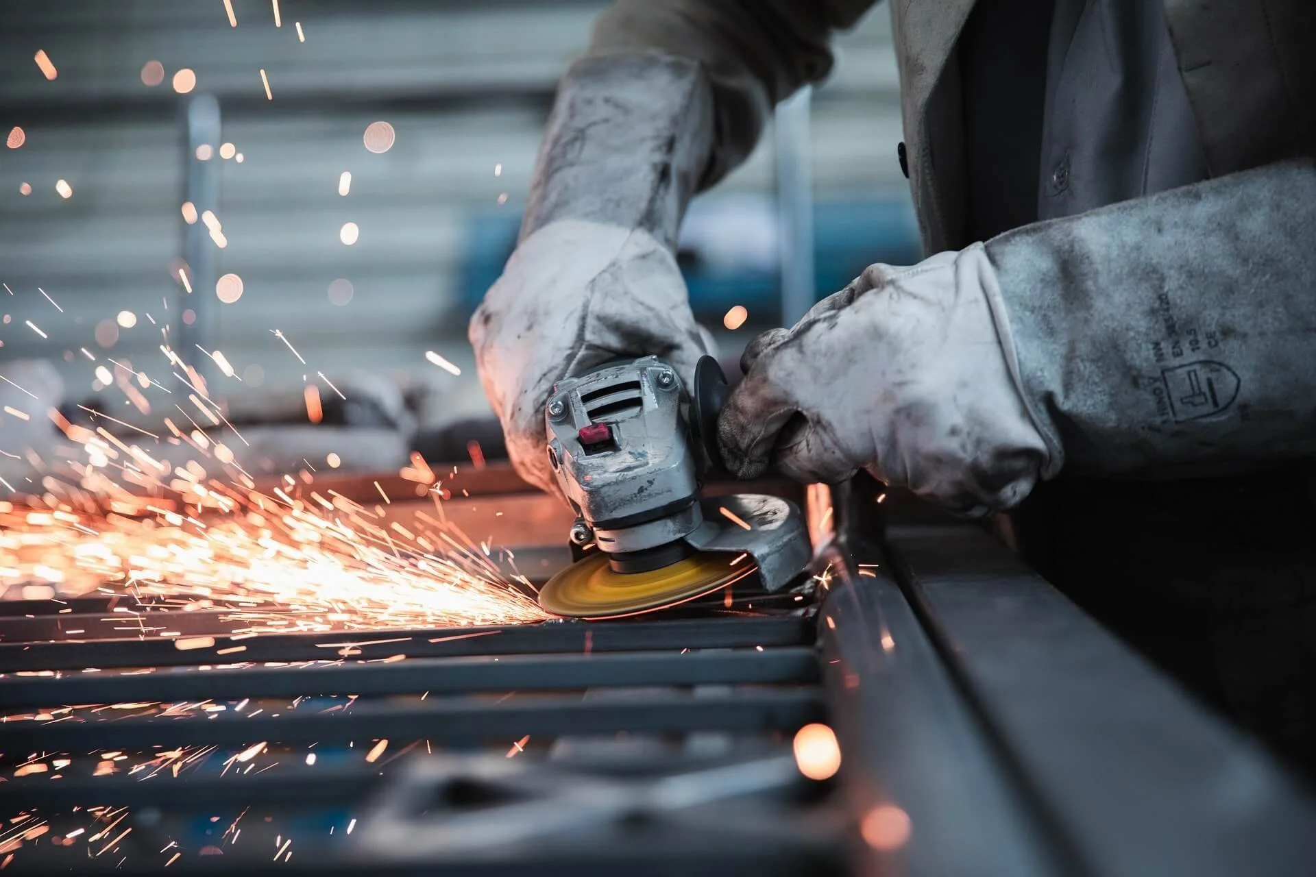 A worker in protective gloves uses a grinding tool to cut metal, creating sparks in an industrial setting.