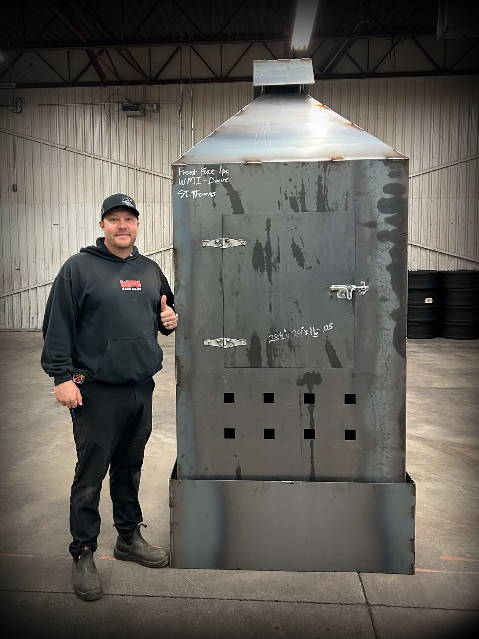 Man wearing black hoodie and cap standing next to a large rusted metal machine in an industrial warehouse.