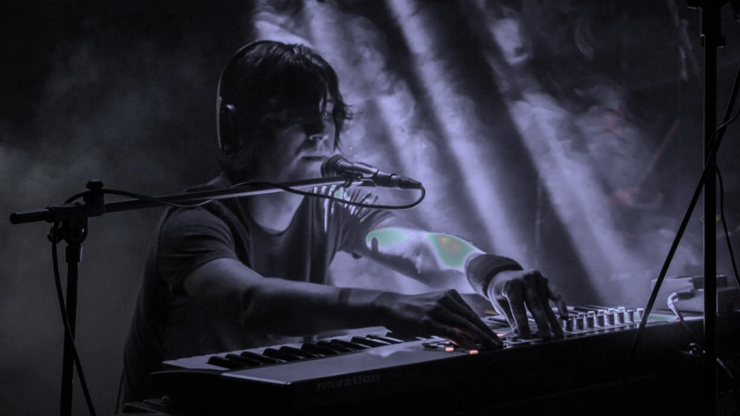Persona tocando el teclado musical y cantando en un micrófono, rodeada de iluminación teatral y humo.