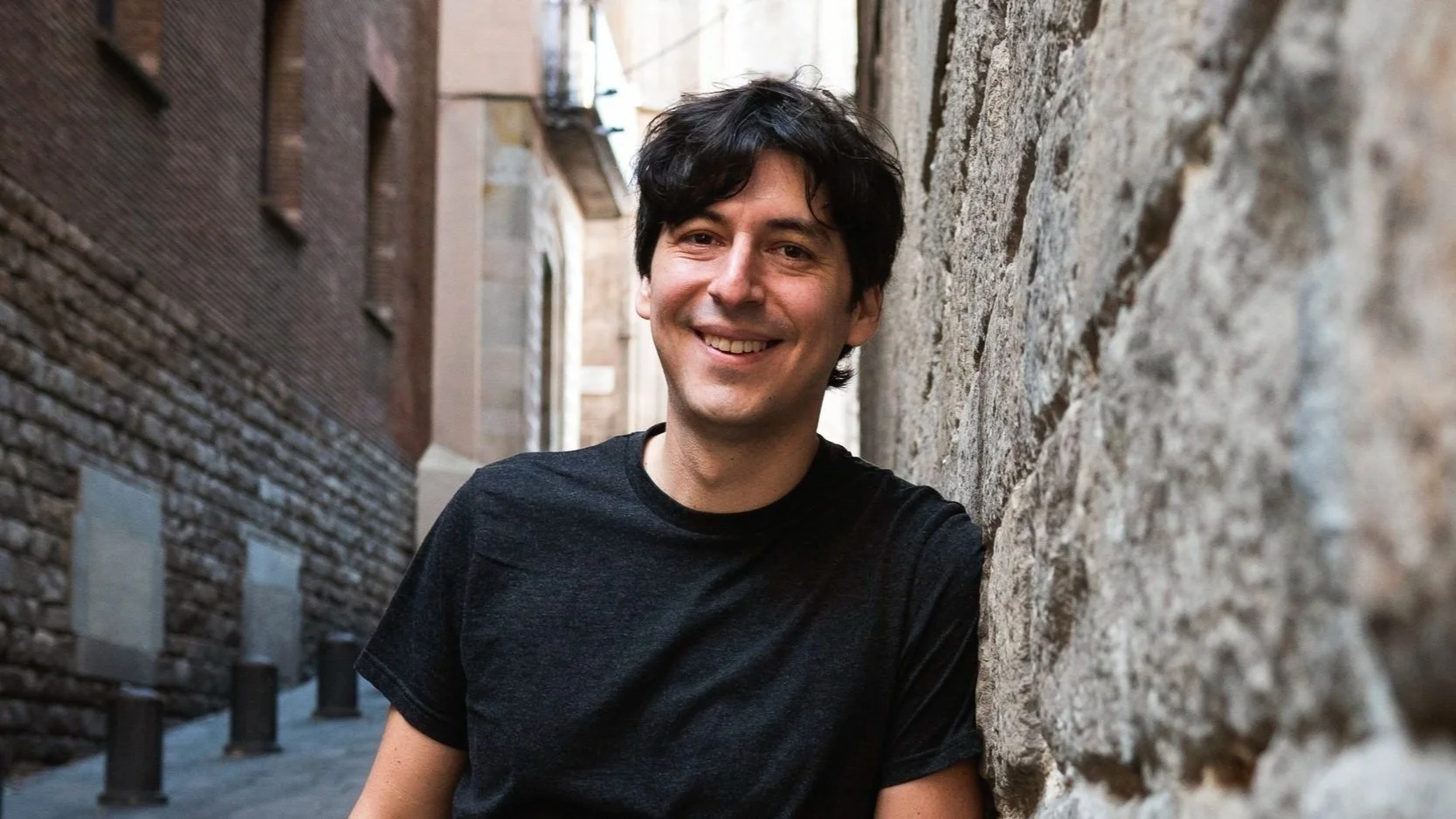 Hombre sonriendo recargado en una pared de ladrillos en un callejón urbano.