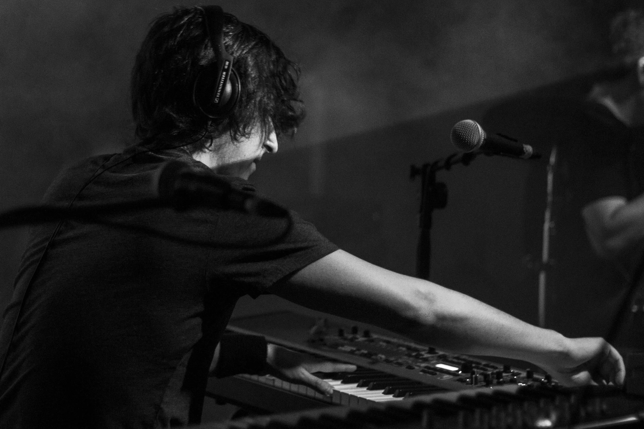 Persona tocando un teclado musical con auriculares puestos, en un escenario con micrófono cercano.