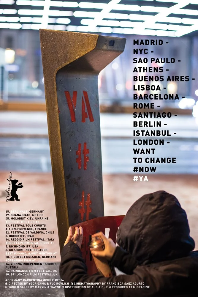 Una persona con capucha interactúa con un poste que tiene la palabra "YA" en letras rojas y un mapa en la parte superior. A la derecha, se enumeran ciudades como Madrid, Nueva York, Sao Paulo, Atenas, Buenos Aires, Lisboa, Barcelona, Roma, Santiago, Berlín, Estambul, Londres. También se leen frases como "WANT TO CHANGE #NOW #YA". Hay textos adicionales sobre festivales de cine y premios en la parte inferior.