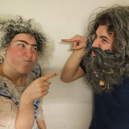 Two people with different hairstyles and beards pointing at each other playfully.