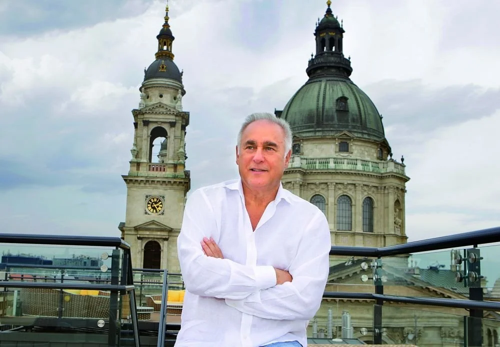 A middle-aged man with gray hair, wearing a white button-up shirt, stands with arms crossed on a rooftop balcony in front of a historic building with a large dome and a clock tower, under a cloudy sky.