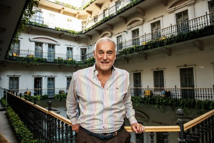 An older man with gray hair and a beard, wearing a striped button-up shirt, smiling and standing on a balcony inside a multi-story hotel courtyard with white walls and black railings, surrounded by plants.